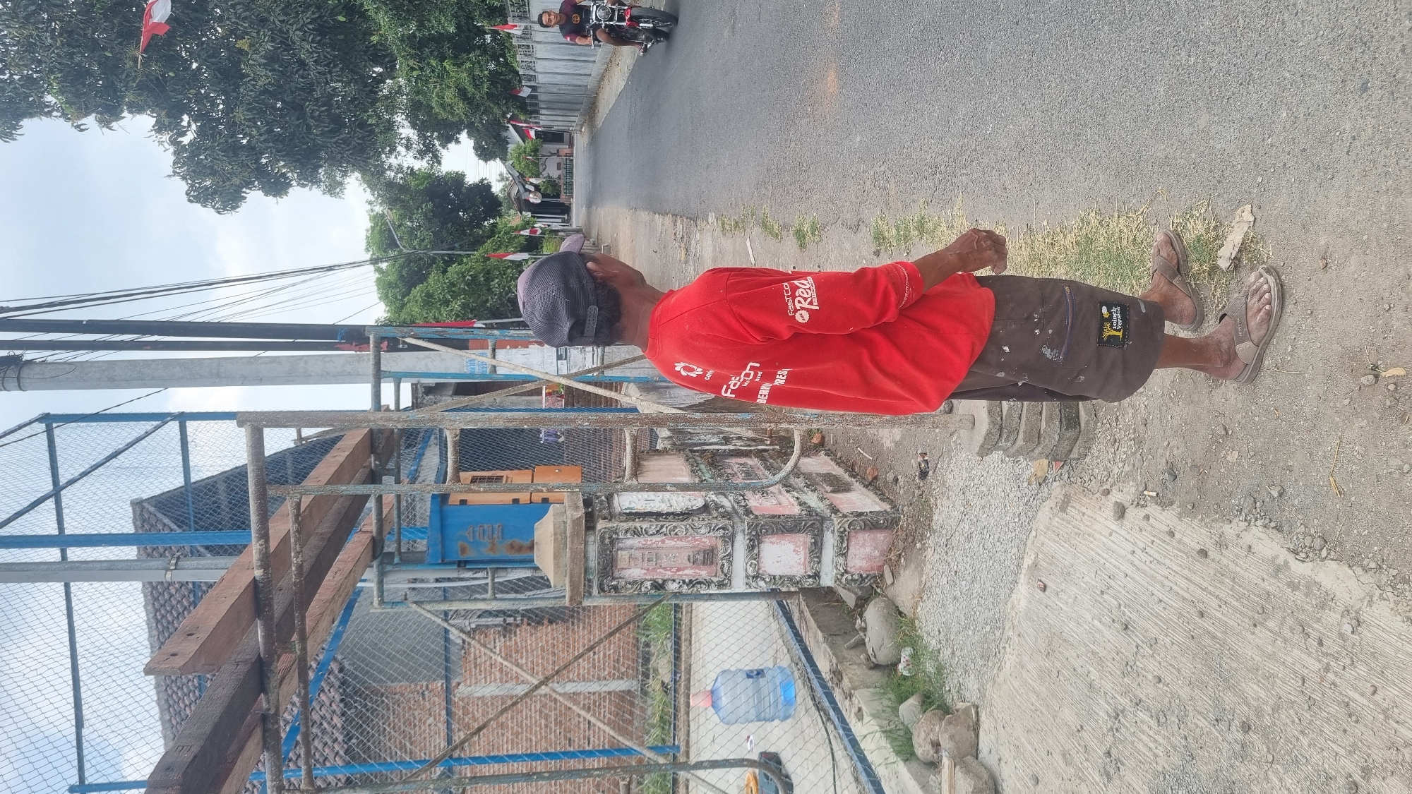A man in a red long-sleeved shirt and brown shorts stands on the side of a paved road in Diwek, Indonesia. He is wearing a dark baseball cap and sandals. The man is looking to his right, towards the road. On his shirt, there is white and red text that appears to be a logo. In the background, a person rides a red motorcycle down the road. To the left of the man, there is a fenced area with a blue structure and a large water jug. The environment appears to be a residential area with houses and trees lining the road. Red and white flags are visible on poles along the street. The sky is overcast.