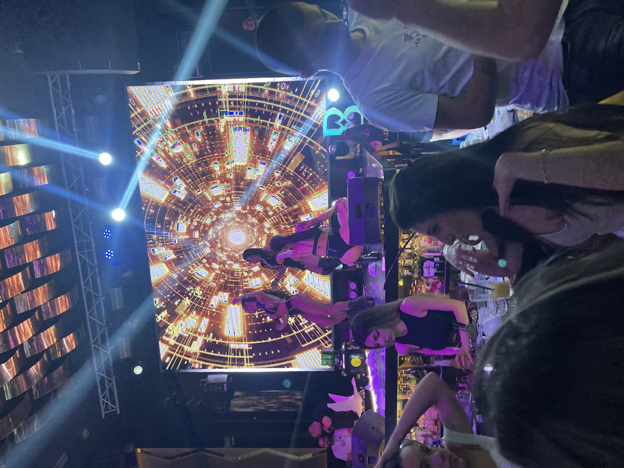 The image shows an interior view of what appears to be a nightclub or bar in Kazanlak, Bulgaria. The predominant colors are purple, gold, and blue. There is a large screen that displays an abstract animated image with concentric circles and bright colors. Two pole dancers are visible in front of the screen. In the foreground, there are multiple people, including a woman with long dark hair and a man wearing a white shirt. There are also cocktail glasses and bottles behind the bar. Stage lighting and decorations contribute to the club's ambiance.