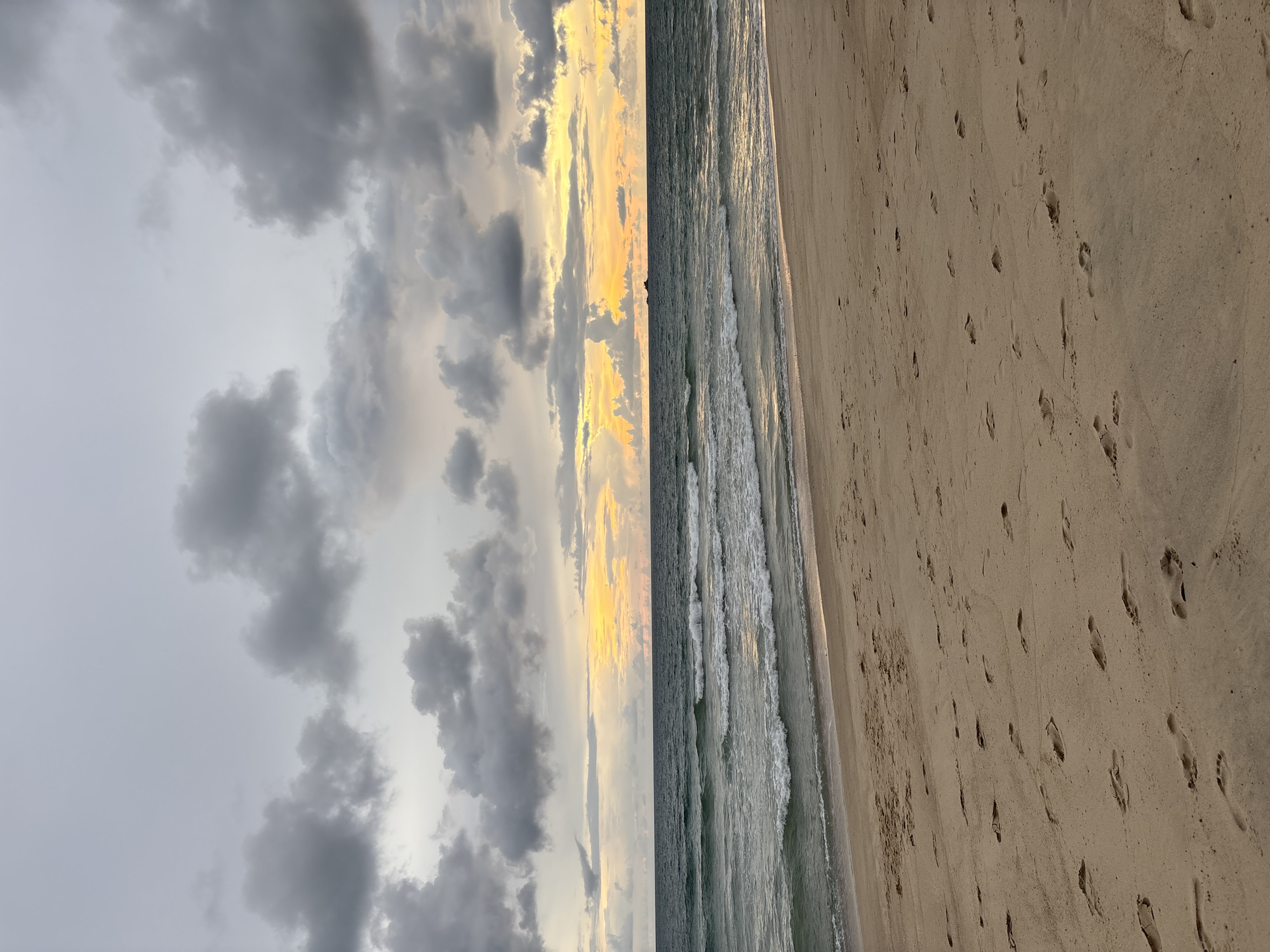 The image depicts a beach scene with a sandy shore covered in footprints. The sea is calm with small waves rolling in, and the water is a deep blue-green. In the distance, a small, dark object, possibly a boat or a rock formation, is visible on the horizon. The sky is overcast with dramatic, dark gray clouds, but there is a band of warm, yellow and orange light breaking through at the horizon, indicating a sunset or sunrise. The overall atmosphere is moody and serene. The location is the beach in Choeng Thale, Thailand, a coastal area known for its picturesque beaches and tropical climate.