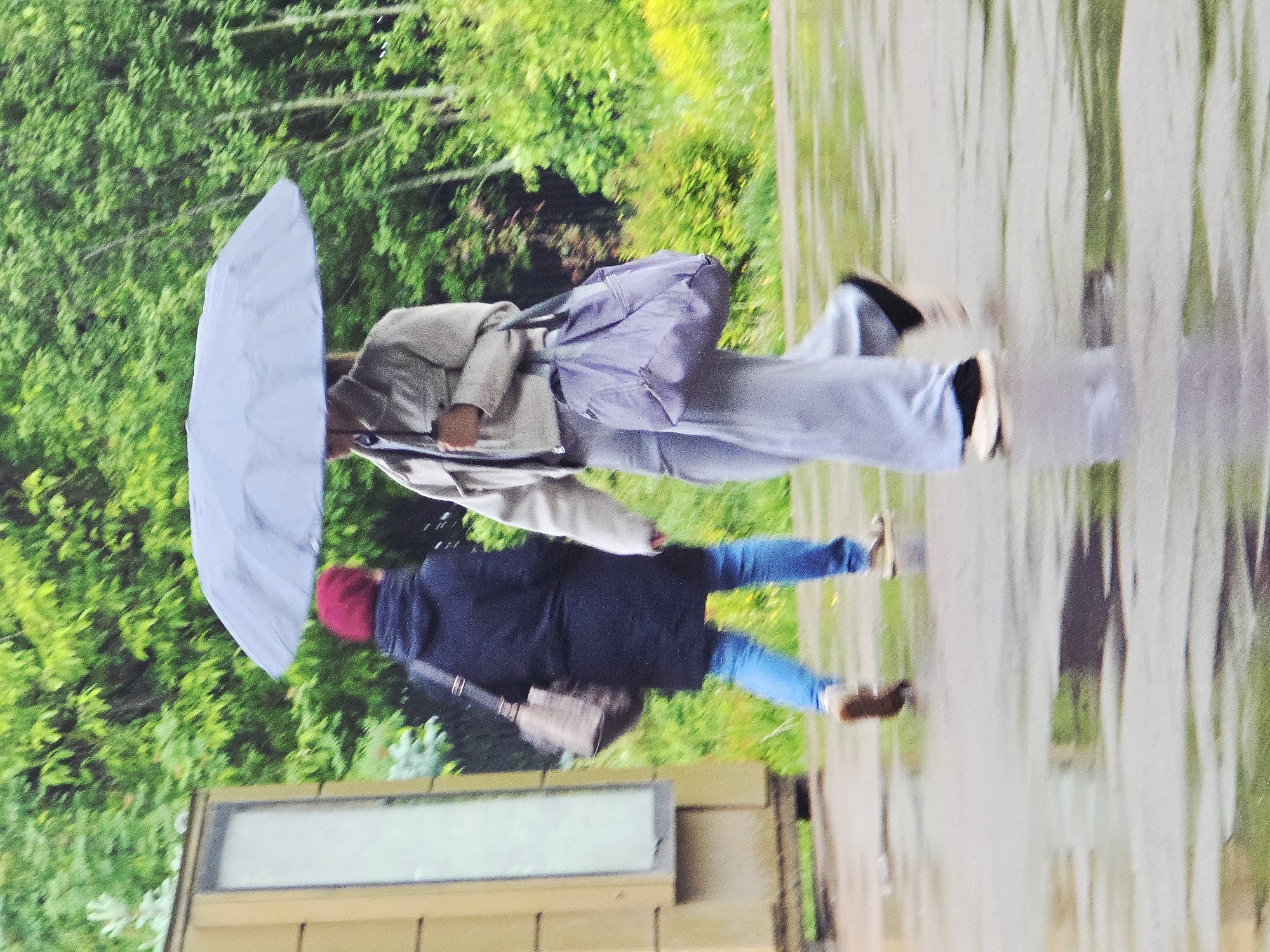 Two people walk on a wet sidewalk beneath an umbrella. One person, wearing a black coat, jeans, and a red hat, and carrying a purse, walks behind another person holding the umbrella and wearing a tan jacket, gray pants, and a purple bag. The setting appears to be outdoors, with trees and a building visible in the background, suggesting a public space. The location might be Moscow, Russia.