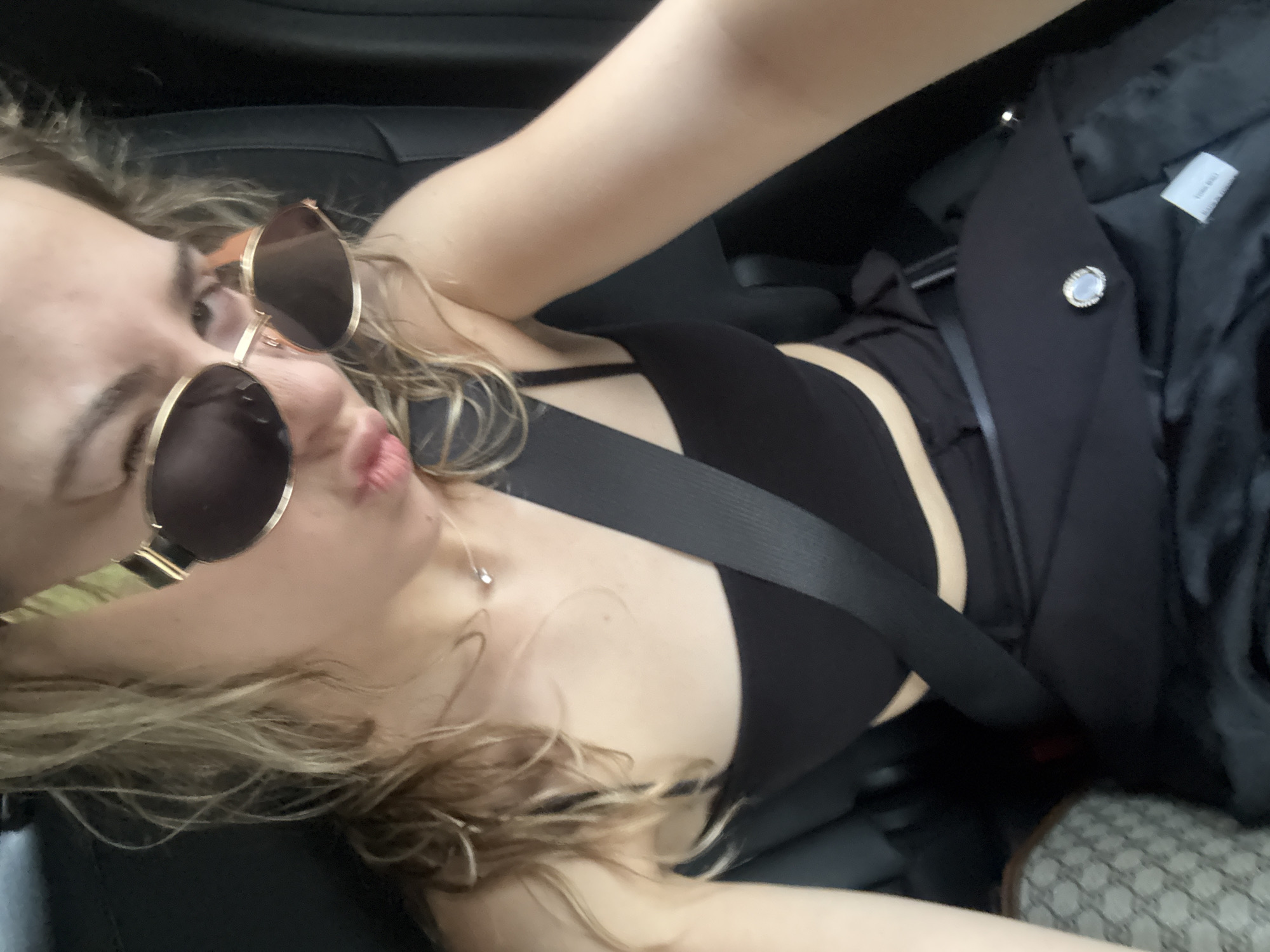 A blonde-haired individual is depicted taking a close-up selfie from the passenger side of a vehicle's rear seat in Sofia, Bulgaria. The individual's face is partially obscured by large, gold-framed sunglasses with dark, oval-shaped lenses, while their lips are pursed in a pout. A thin silver chain necklace with a small, clear stone pendant is visible around their neck. They are wearing a black, strapless or thin-strapped crop top. A black diagonal seatbelt crosses their chest and lap. A black garment, possibly a jacket or blazer, with a prominent silver and white circular button or emblem is draped across their lap. A white manufacturing tag with black text is visible on the inside lining of this garment. In the lower left quadrant of the image, a beige or cream-colored bag with a repeating patterned monogram is partially visible, resting next to the individual's side. The vehicle's interior features dark upholstery on the seats and headrests.