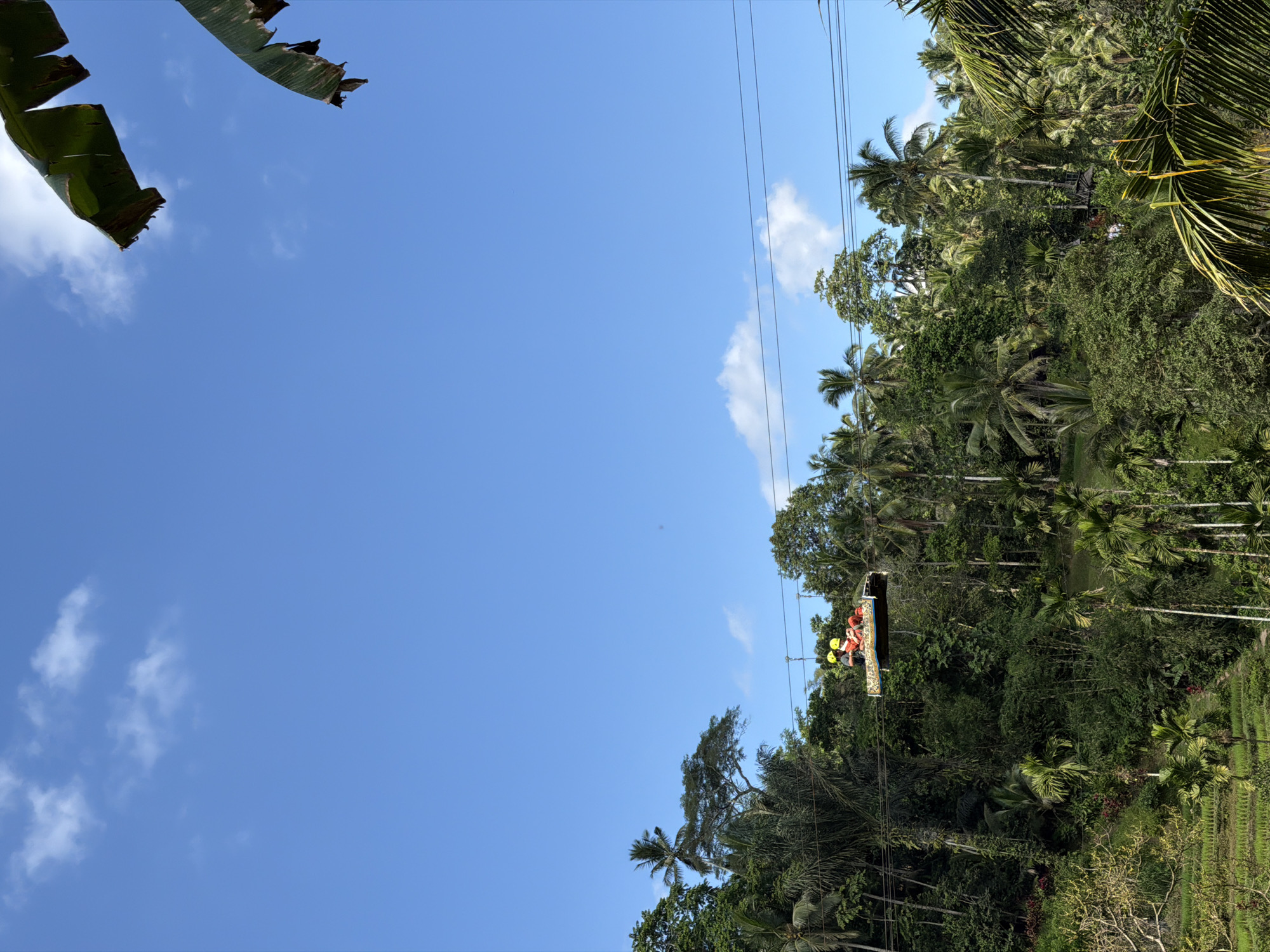Two individuals wearing yellow helmets are seated on a raised platform, suspended from wires, as they traverse across a lush, tropical landscape. The platform, which appears to be a vibrant red cushion with a yellow trim, is positioned against a backdrop of dense green foliage and tall palm trees. The individuals are positioned side-by-side, with one person appearing to be looking forward and the other looking slightly to the side. The scene is set under a clear blue sky with scattered white clouds. Below the platform, terraced rice paddies are visible, contributing to the vibrant green hues of the environment. The location is identified as Tegallalang, Indonesia, suggesting an aerial perspective of the famous rice terraces and surrounding jungle.