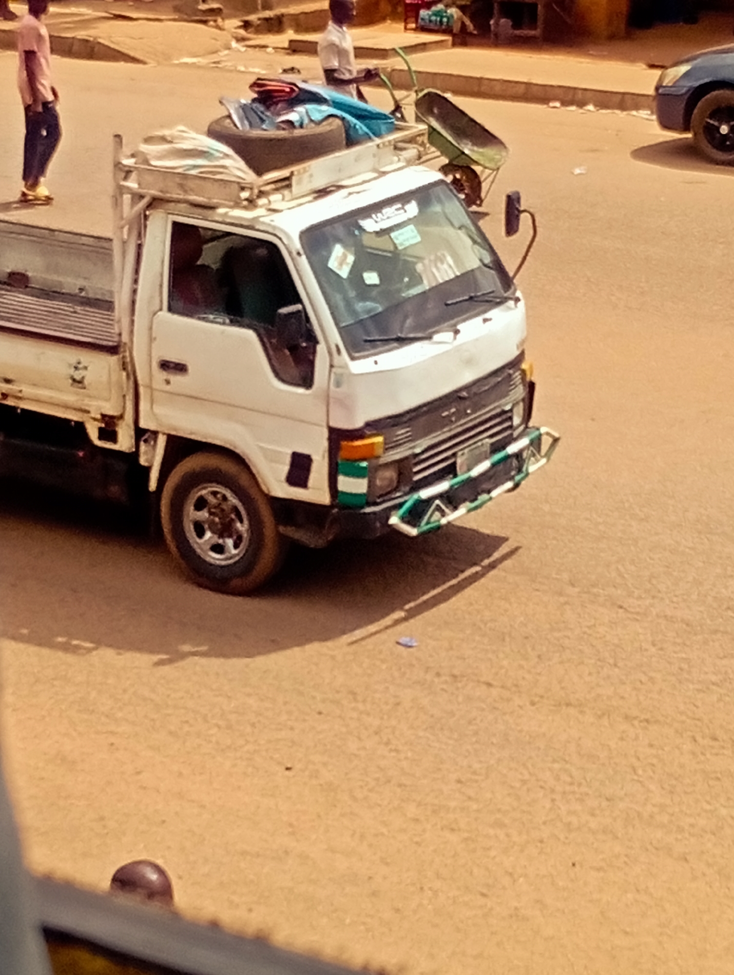 The image shows a white pickup truck parked on a dusty, unpaved road in what appears to be a market or busy street area in Bukuru, Nigeria. The truck bed is loaded with various items, including what looks like a spare tire, fabric bundles, and possibly tools. A wheelbarrow is also secured to the top of the truck.
Two people are visible in the background. One, a man wearing a light pink shirt and dark pants, stands to the left of the truck. Another man, wearing a white shirt, stands further back on the right. The scene suggests an active environment, possibly a transportation hub or a commercial area where goods are being moved.
The truck has a custom bumper with green and white stripes. There is some visible text on the windshield of the truck, though it is not clearly legible. The lighting indicates it is daytime, with the sun casting distinct shadows, suggesting a sunny day. The overall impression is a scene of everyday commerce and transportation in a Nigerian town.