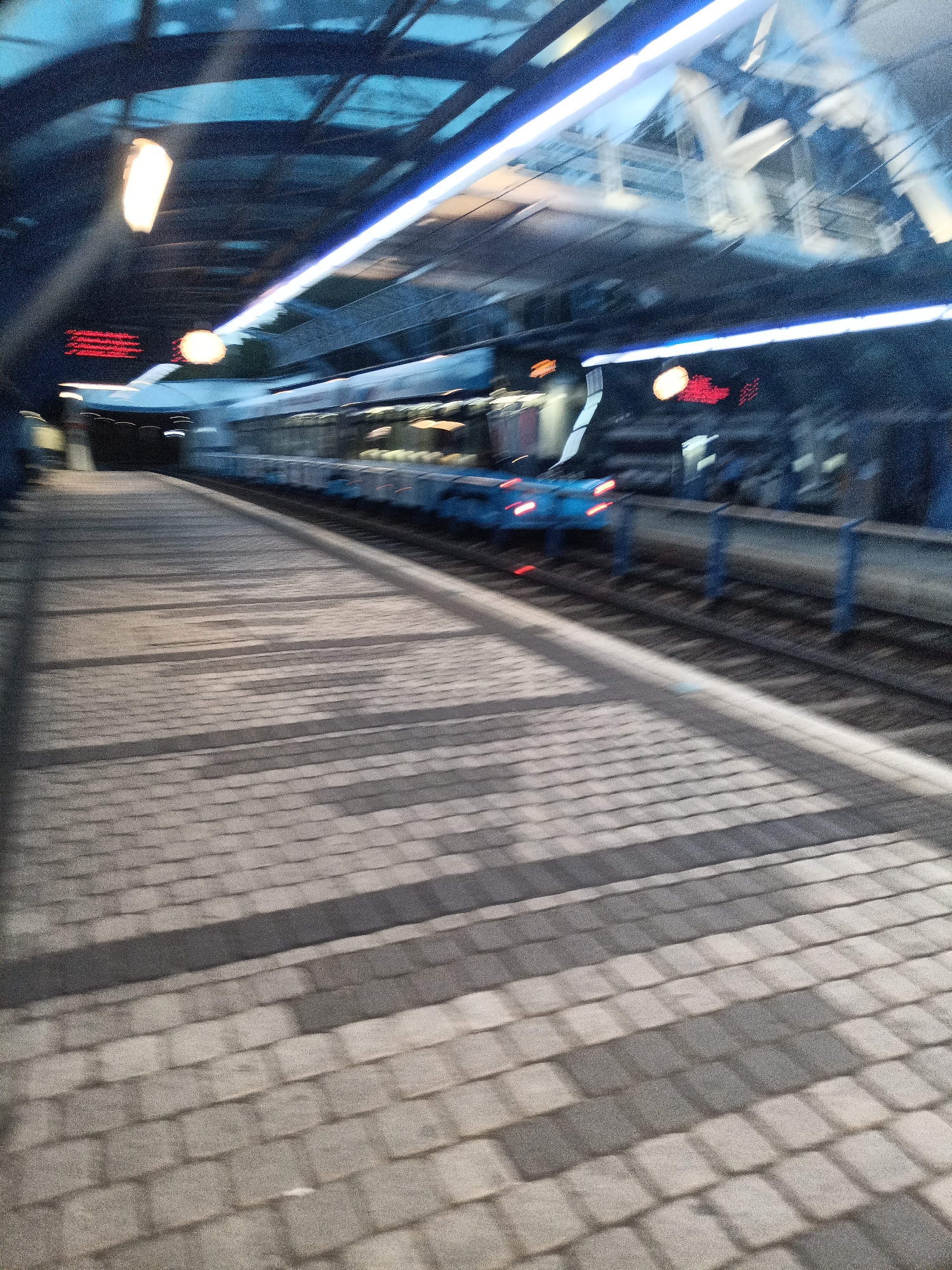 An out-of-focus, low-light nighttime exterior view shows a train at a covered platform. The train, which appears to be blue, is stopped at the station, with its red taillights visible. The platform is paved with rectangular stones, creating a striped pattern of light and shadow. Above the platform, a blue translucent roof is supported by metal beams. Bright white and blue lights line the underside of the roof. A red digital sign is visible on the building behind the train. The scene is likely a train station in Prague, Czechia, given the context.