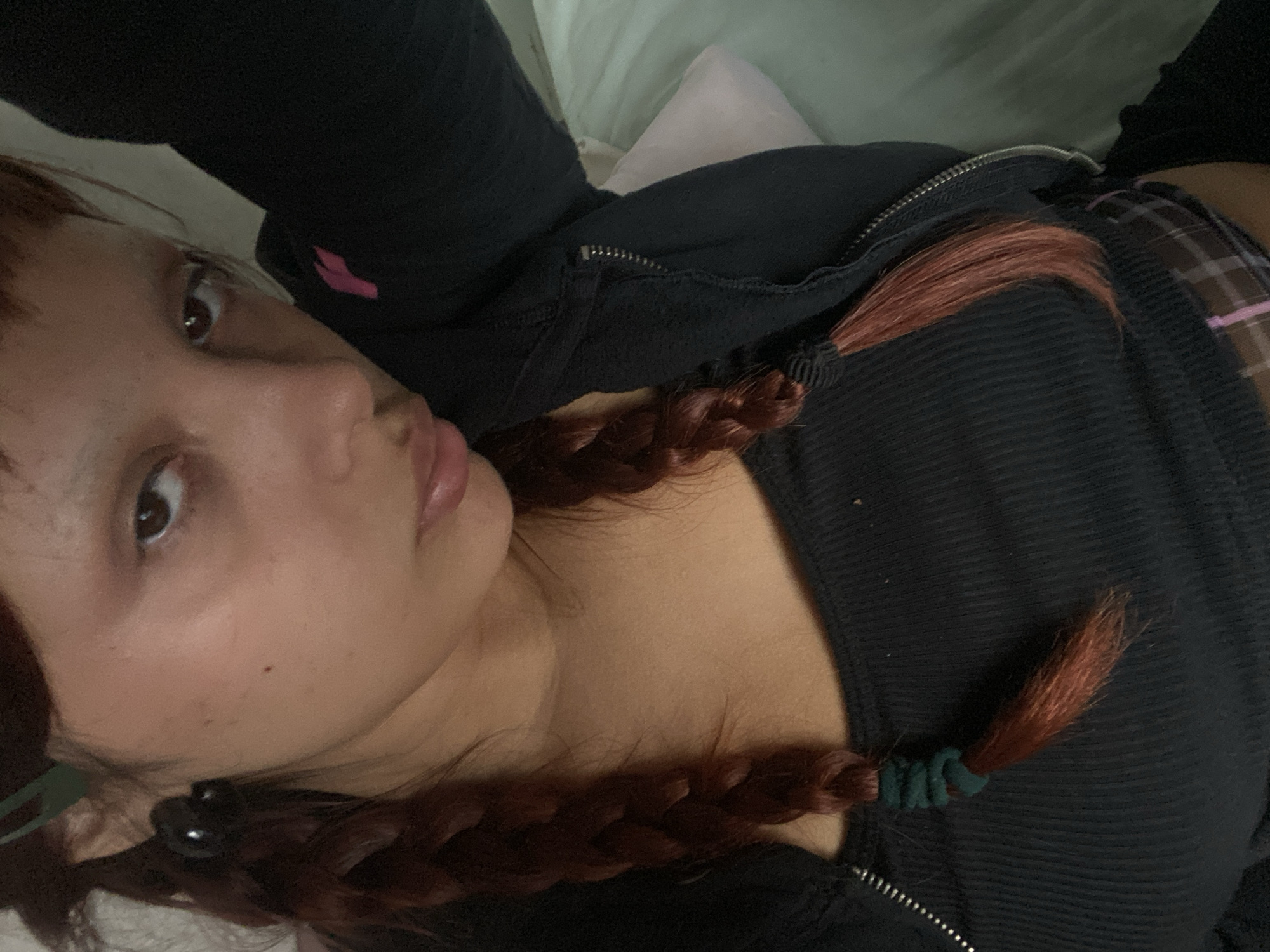 The image shows a person lying on a light-colored surface, possibly a bed or cushion. The person has reddish-brown hair braided into pigtails secured with dark-colored elastic bands. They are wearing a dark-colored, zip-up, ribbed shirt or jacket. The person's face is visible, looking towards the camera with an expressive gaze. The background includes a pink pillow or cushion. The image provides no data to determine a location in Buenos Aires, Argentina, or any other city.