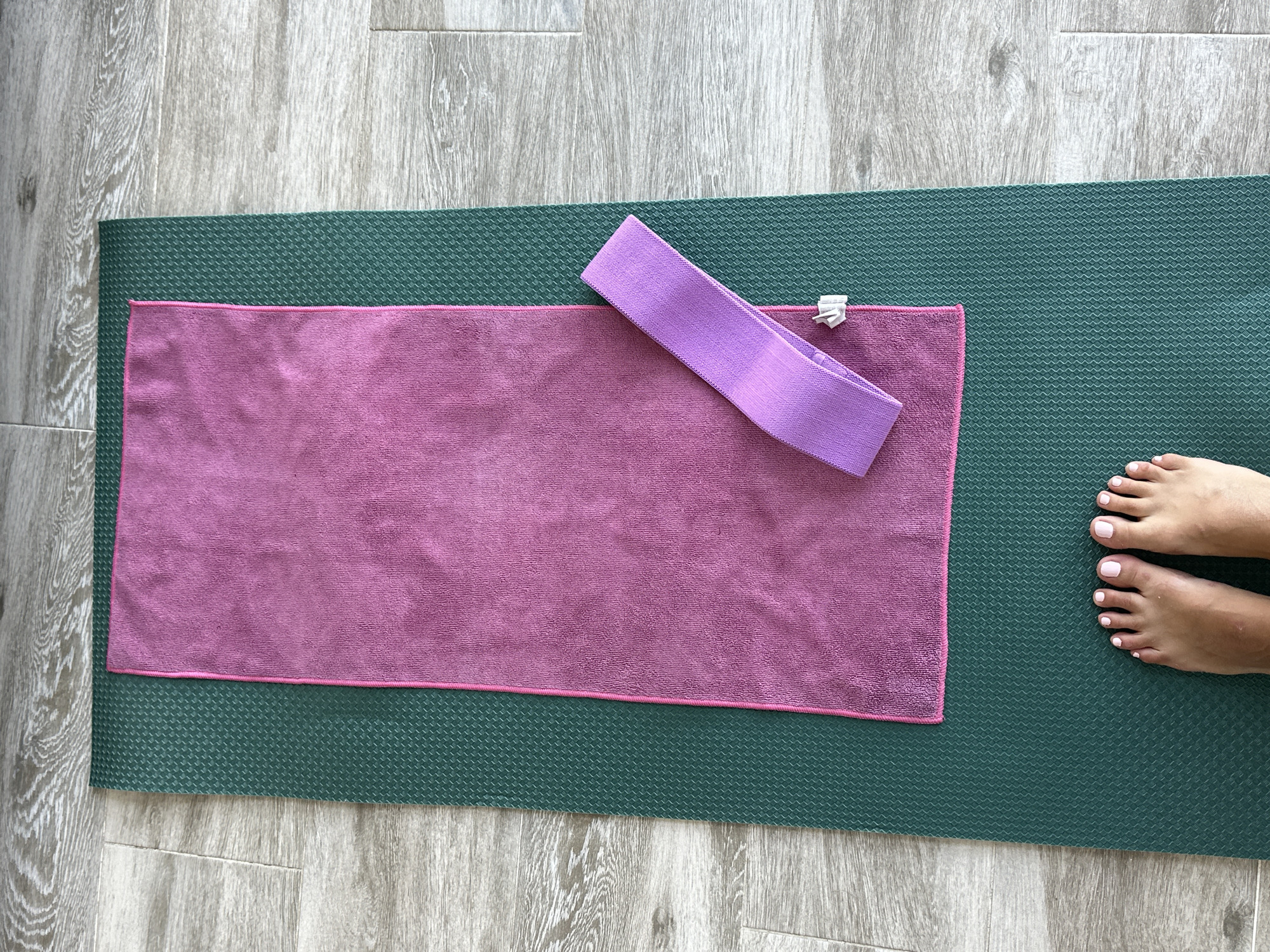 An overhead view shows a dark green yoga mat on a light wood-grain floor. Centered on the yoga mat is a pink microfiber towel. To the right of the towel, on top of the towel, is a purple resistance band. The bottom of the frame shows a pair of feet with white-painted toenails standing on the yoga mat. The scene is set in a domestic environment, likely a home gym or bedroom, in Torremolinos, Spain.