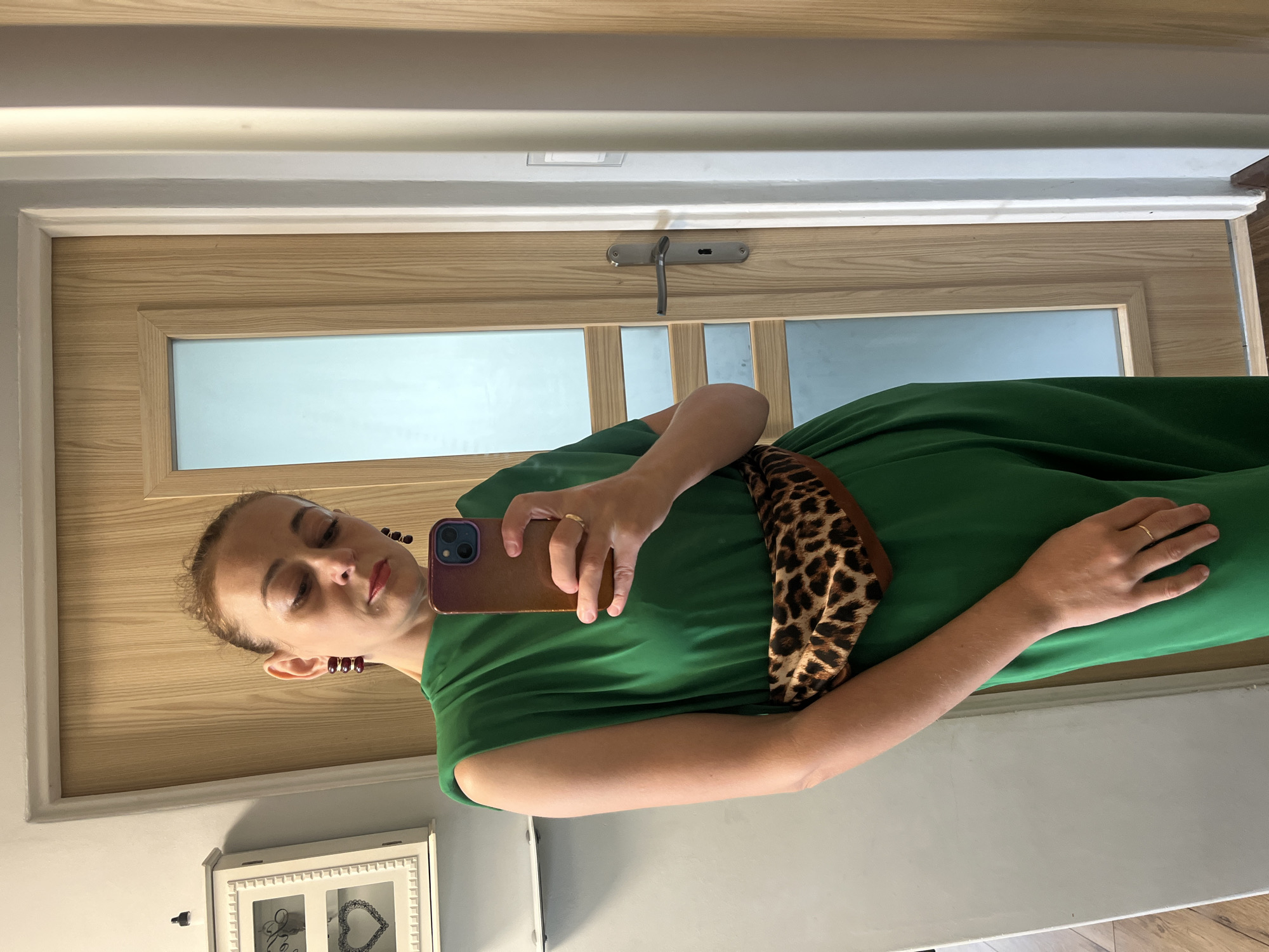 A woman stands in a room, taking a selfie in a mirror. She is wearing a green dress with a leopard print belt and large red earrings. She holds a smartphone in her right hand, with her left hand resting on her hip. The background shows a wooden door with a glass pane, and a white wall with a decorative cabinet. The flooring appears to be wood. The lighting suggests it is daytime, possibly indoors near a window.
