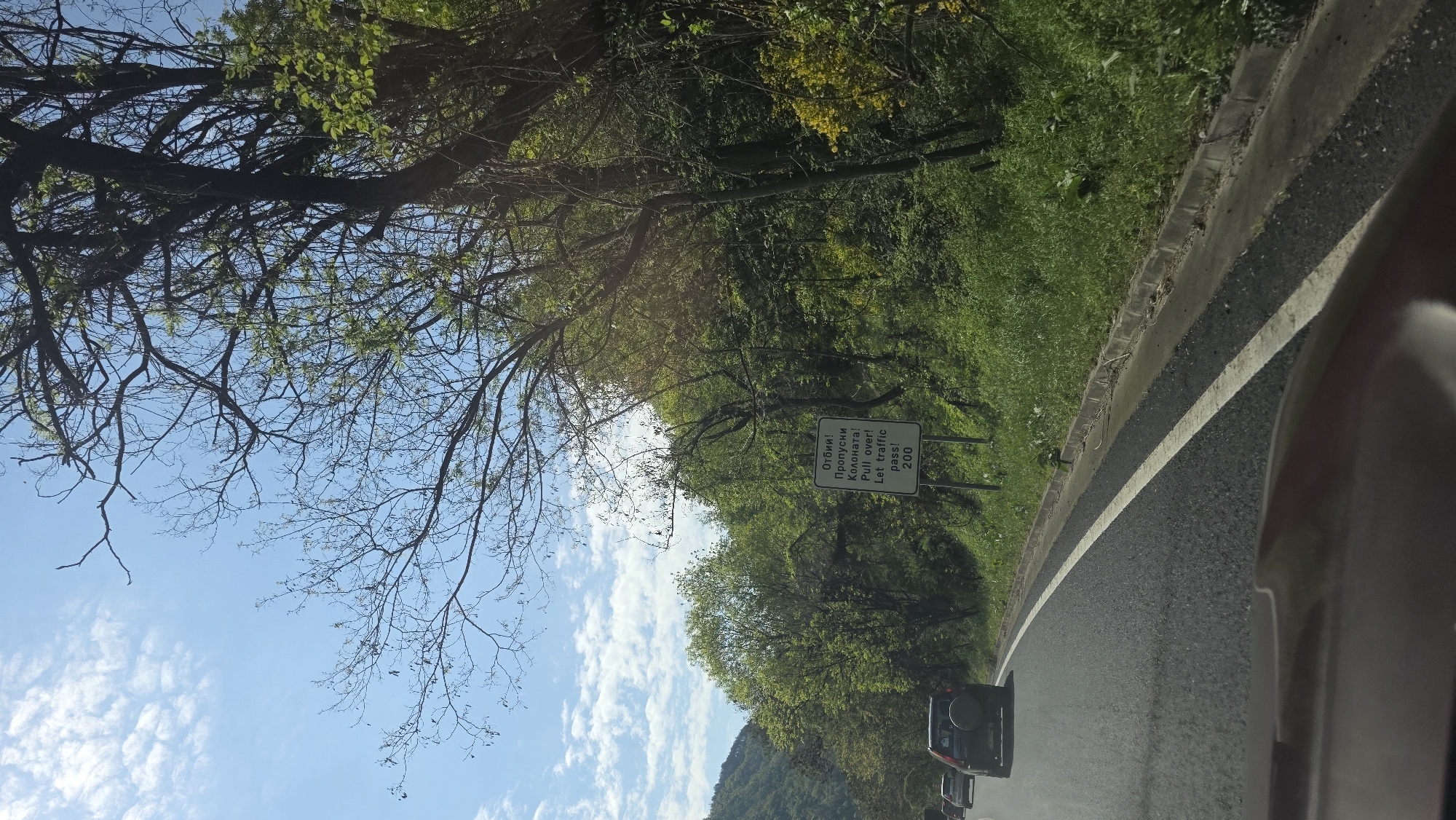 The image depicts a road in Stara Kresna, Bulgaria, lined with trees and vegetation. A road sign is visible, featuring text in both Bulgarian and English: "Pull over! Let traffic pass! 200". Several cars are driving on the road. A portion of a vehicle's interior is visible in the lower right corner. The sky is blue with scattered clouds, and tree branches are visible overhead.