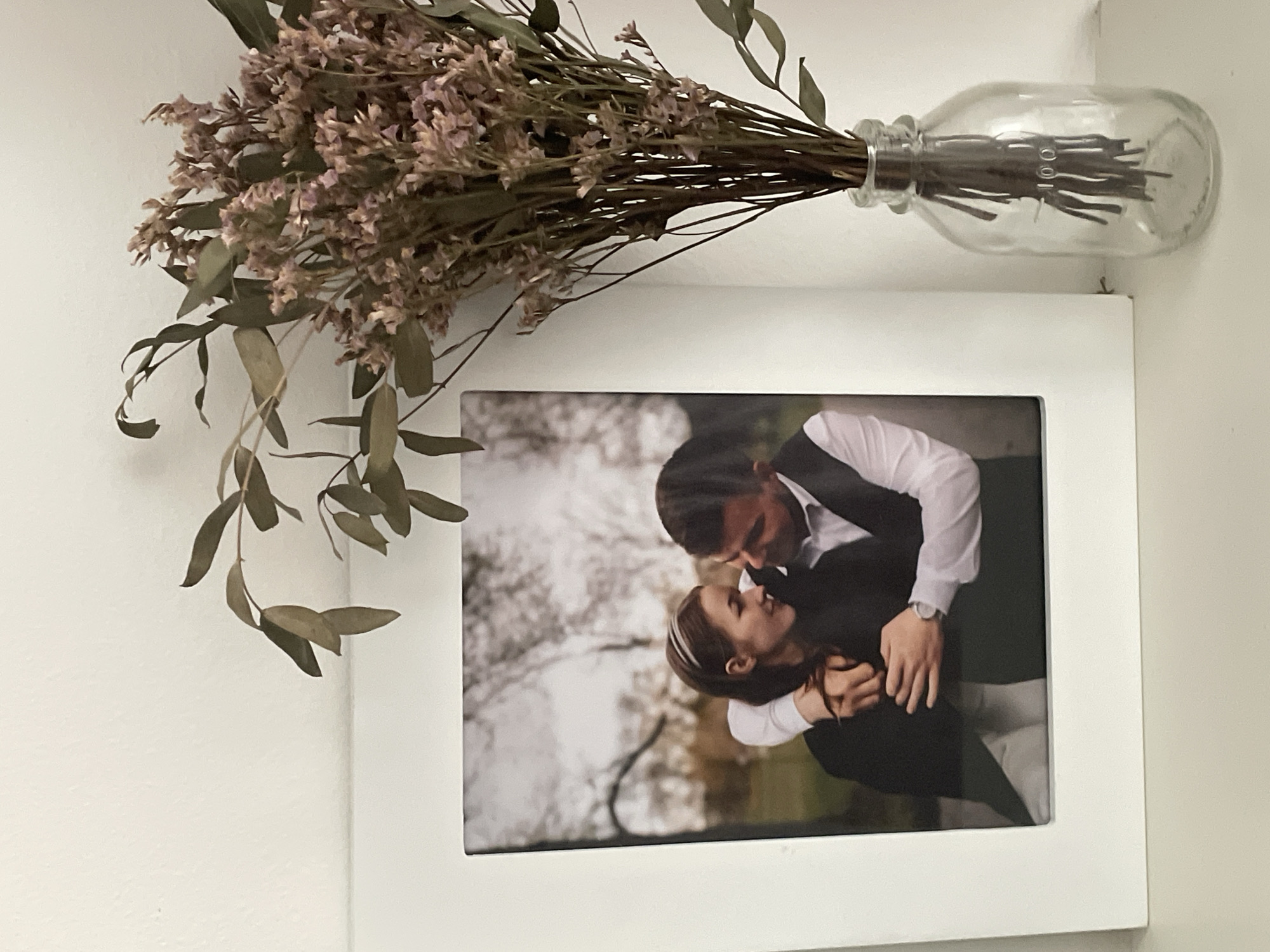 A white picture frame holds a black and white photograph of a man and woman embracing outdoors, with trees in the background. The woman, wearing a dark coat and a headband, is looking at the man, who is dressed in a white shirt and vest. His arms are around her. To the right of the frame, a clear glass bottle with a "100" marking contains a bouquet of dried flowers and eucalyptus branches. The objects are displayed on a white surface, against a plain white wall. The scene is indoors, likely in a home environment. The city of Nidda, Germany is not depicted in the image.