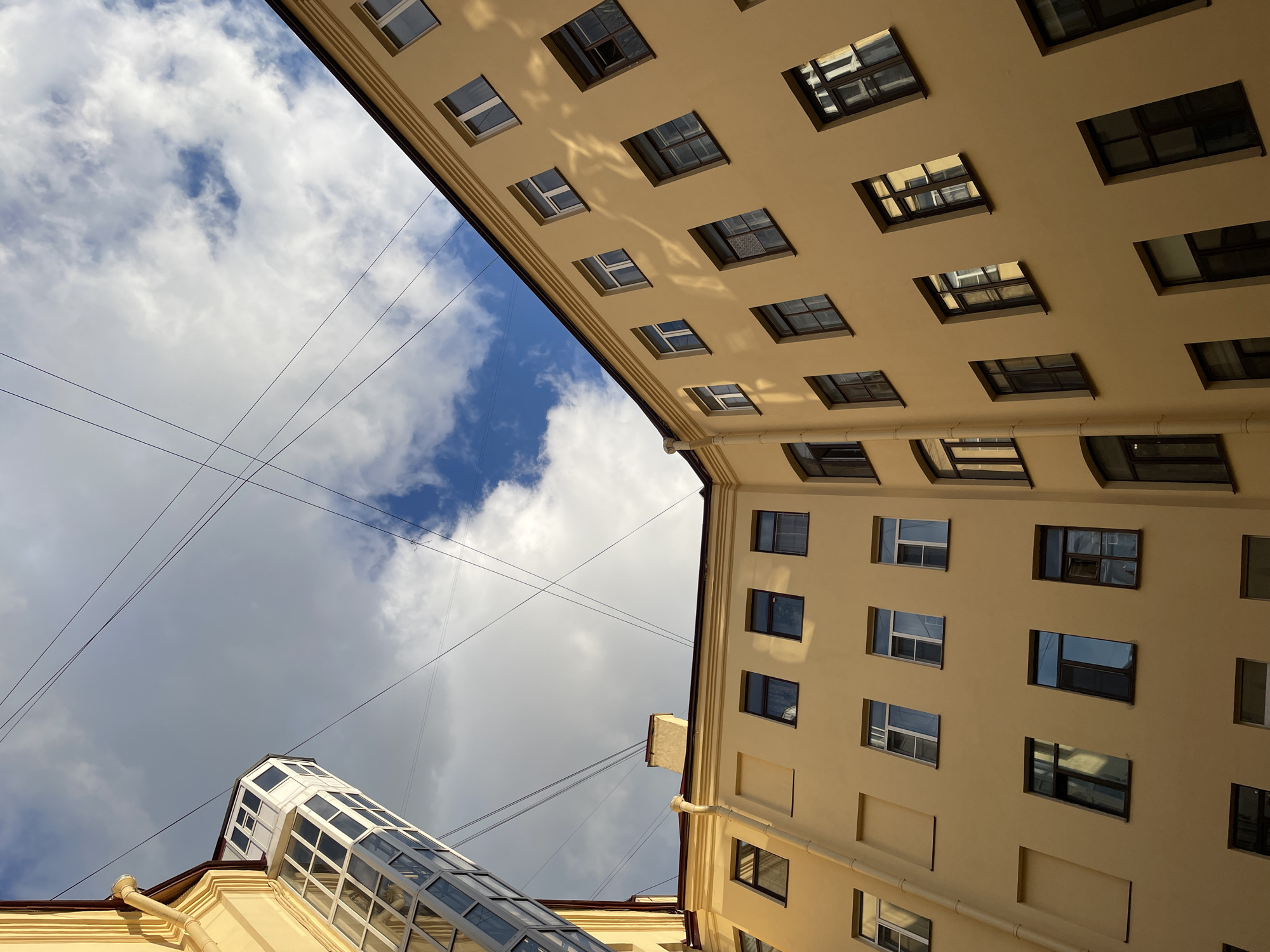 The image captures an upward view of the facade of a pale yellow building in Saint Petersburg, Russia. The architecture appears to be historic, with multiple windows visible on the upper floors. A modern glass elevator shaft is attached to the corner of the building on the left, contrasting with the older structure. Above, the sky is filled with a mix of white clouds and patches of blue, with thin dark wires crisscrossing the frame. The lighting suggests it might be late afternoon or early morning, with shadows cast on the building's surface. There are no people or animals visible in the scene. The overall impression is of an urban architectural detail, typical of a courtyard or narrow street in the historic center of Saint Petersburg.