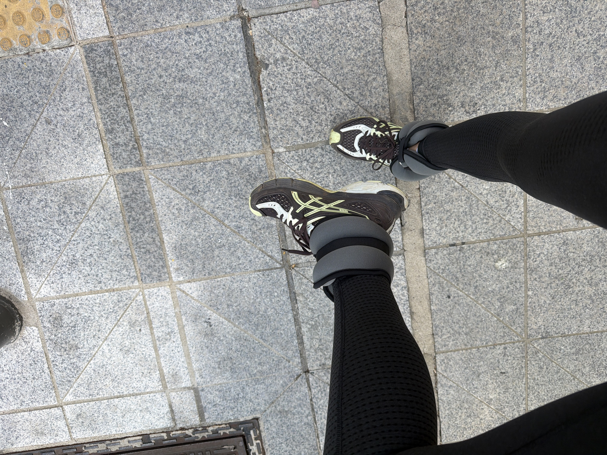 The image shows a top-down view of a person's legs and feet standing on a tiled pavement. The person is wearing black athletic leggings and athletic shoes with ankle weights. The pavement is composed of grey square tiles. In the lower left corner of the frame, a portion of a manhole cover is visible. The location is identified as Seoul, South Korea, based on the pavement design.