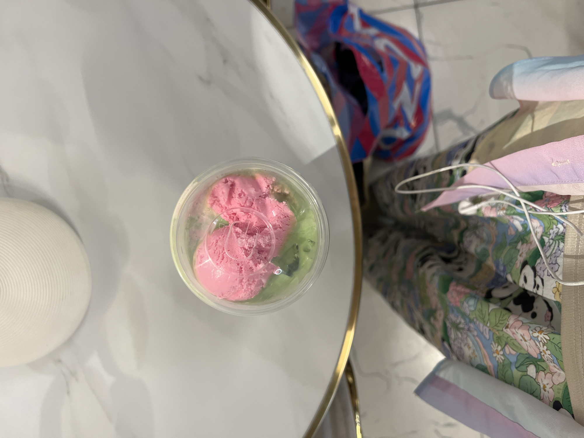 The image is an overhead shot of a clear plastic cup containing two scoops of pink ice cream on top of a green substance, likely more ice cream or a sorbet. The cup is placed on a white marble-like surface, possibly a table, with a gold trim. In the background, there is a large, textured white object and a colorful striped bag. In the foreground, a person's legs are visible, wearing patterned pants and a light-colored garment. The overall setting appears to be indoors, potentially a cafe or restaurant, though no specific landmarks in Almaty, Kazakhstan are discernible from the image. The lighting suggests daytime.