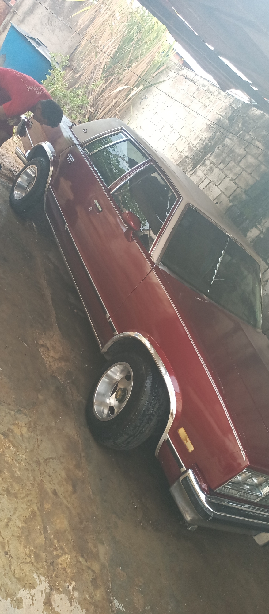 A person wearing a red shirt is washing a maroon car in what appears to be an outdoor, covered area, possibly a garage or carport. The car is a vintage model with chrome accents and a tan vinyl top. The ground is wet and muddy, suggesting it has been raining or the car is being washed. Tall, dry grass and a brick wall are visible in the background, along with a blue plastic object. Sunlight streams in from the side, creating bright highlights on the car and casting shadows. The scene appears to be taking place during the daytime. The person's face is not visible, but their posture suggests they are focused on cleaning the car. There is no visible text in the image.