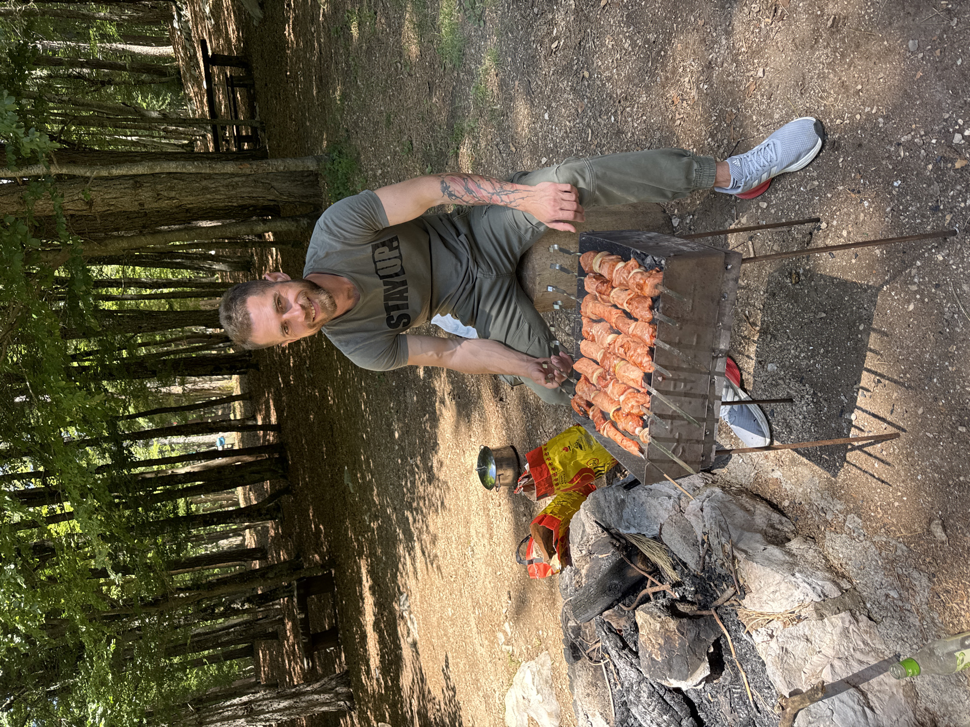 The image depicts an outdoor scene in a sunlit forest clearing in Mali Bostur, Montenegro. An adult male is seated on a wooden log, facing the camera with a smile, while grilling meat skewers on a rectangular metal grill. He wears a dark olive green t-shirt with "STAY UP" printed in capital letters, matching olive green cargo pants, and light grey Adidas athletic shoes with red accents. A multi-colored tattoo covers his left forearm. Approximately ten skewers with raw meat and onion pieces are visible on the grill, positioned over coals. To the immediate left of the grill, a stone-lined fire pit contains charred wood and ash. Behind the fire pit are two yellow and red patterned bags, one partially open, and a silver pot with a lid resting on the ground. A green glass bottle is partially visible in the bottom left foreground. The ground consists of dry earth, scattered fallen leaves, and small rocks, with dappled sunlight and shadows cast by numerous tall trees with green foliage. In the background, two wooden picnic tables with benches are visible under the tree canopy.