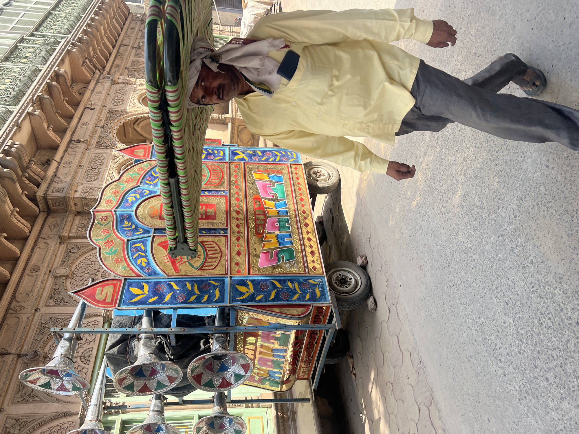 The image captures a man standing next to a brightly decorated vehicle, likely a rickshaw or small cart, in Vrindavan, India. The vehicle features vibrant colors and ornate designs, including floral patterns, gold accents, and the word "SHAKTI" written in colorful block letters. Below the main body of the vehicle, there are several reflective, conical ornaments hanging. The man is wearing a light yellow button-down shirt, dark gray pants, and sandals. He also has a white cloth wrapped around his head. The background shows a building with intricate architectural details, suggesting a historical or religious site. The scene is set on a paved street or sidewalk.