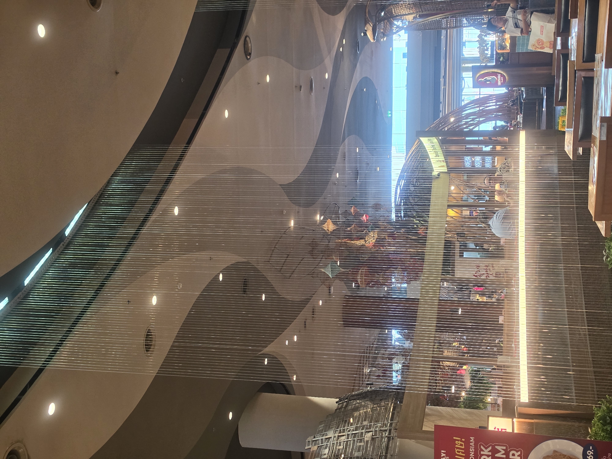 This image shows the interior of a shopping mall in Bangkok, Thailand. The photo appears to be taken from a low angle, looking upward. The main architectural features include a layered, curved ceiling with embedded lights and what seems like a decorative light installation of numerous thin, vertical strands hanging from the ceiling. A cafe or restaurant area with tables and chairs can be seen on the right side of the frame. People are present in the restaurant area, and there are also visible signs with Thai script, confirming its location.