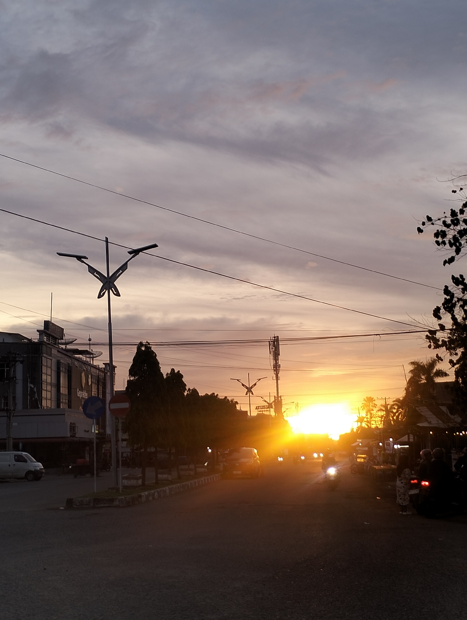 The photograph captures a street scene at sunset in Lhokseumawe, Indonesia. The sky is filled with a mix of gray and orange hues as the sun sets in the distance, casting a warm glow over the area.
The street is lined with buildings on the left and trees on the right. A white van is parked on the side of the road near a traffic sign indicating a one-way street. The road surface is dark, and the scene is generally dimly lit due to the sunset.
Several vehicles, including cars and motorcycles, are visible on the road, their headlights creating streaks of light. On the right side of the street, people are gathered near what appears to be street food stalls.