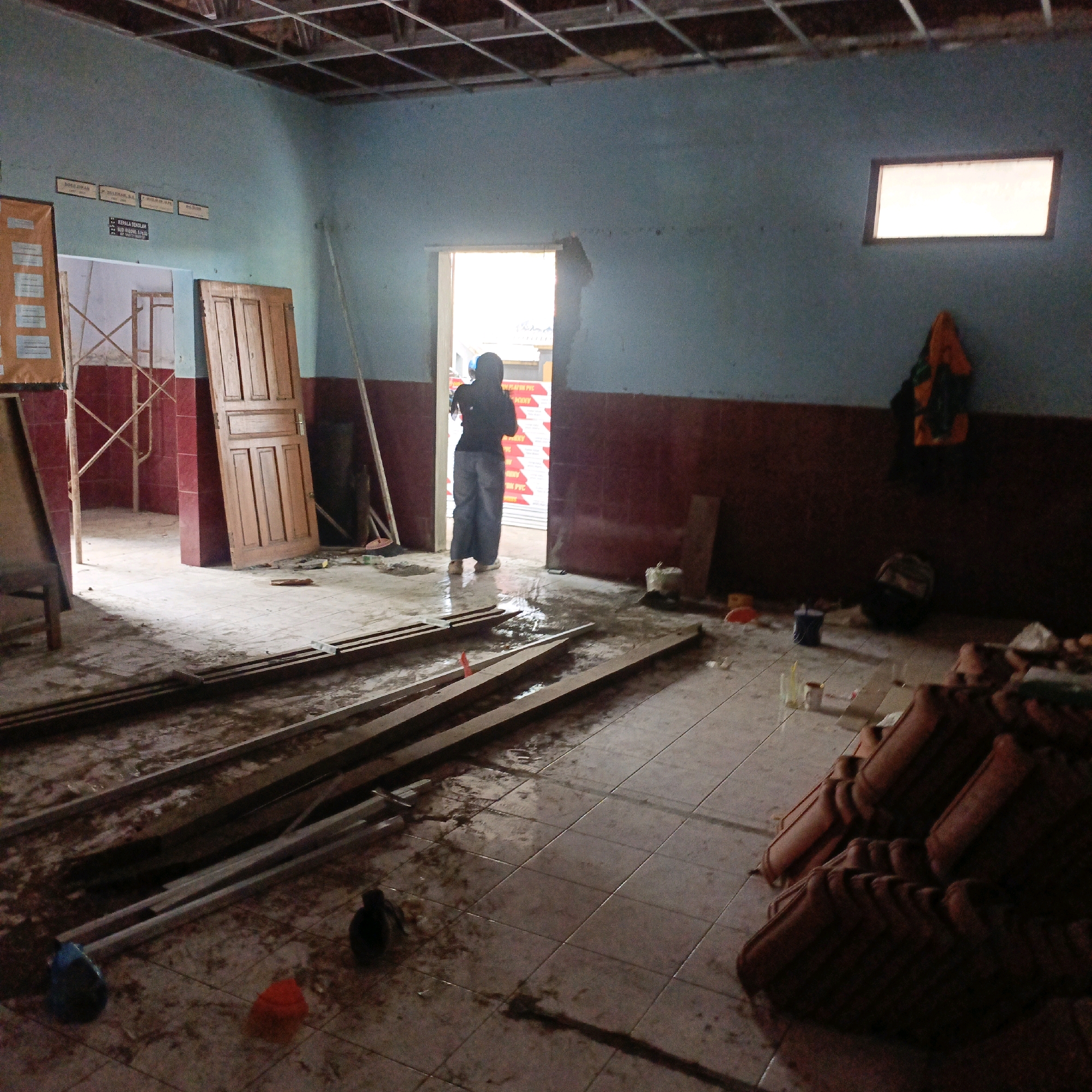 The image depicts an interior scene, possibly a room under renovation or construction. The walls are painted a light blue on the upper half and a darker reddish-brown on the lower half. The ceiling appears to be a suspended grid system, with exposed wiring or structure. The floor is tiled and shows signs of water or dampness, with scattered debris and materials.
In the center of the room, a doorway leads to a brighter area, presumably outside or another section of the building. A person wearing a head covering and dark clothing, with light-colored pants and shoes, is walking through this doorway.
Various construction or renovation materials are present, including wooden planks laid out on the floor, stacks of what appear to be roofing tiles or similar building materials in the foreground right, and what looks like scaffolding in the background left. There are also a few scattered objects like buckets, bags, and possibly some discarded containers.
A wooden door stands open to the left of the central doorway. A small rectangular window is visible high on the right wall, illuminated from within. A piece of orange and green fabric, possibly clothing, is hanging on the wall to the right of the window.
The image captures a moment in a functional space that is currently in a state of disarray due to ongoing work. The lighting suggests it's daytime, with light coming in from the doorway and the window. The overall impression is one of a work-in-progress within an institutional or residential building.
