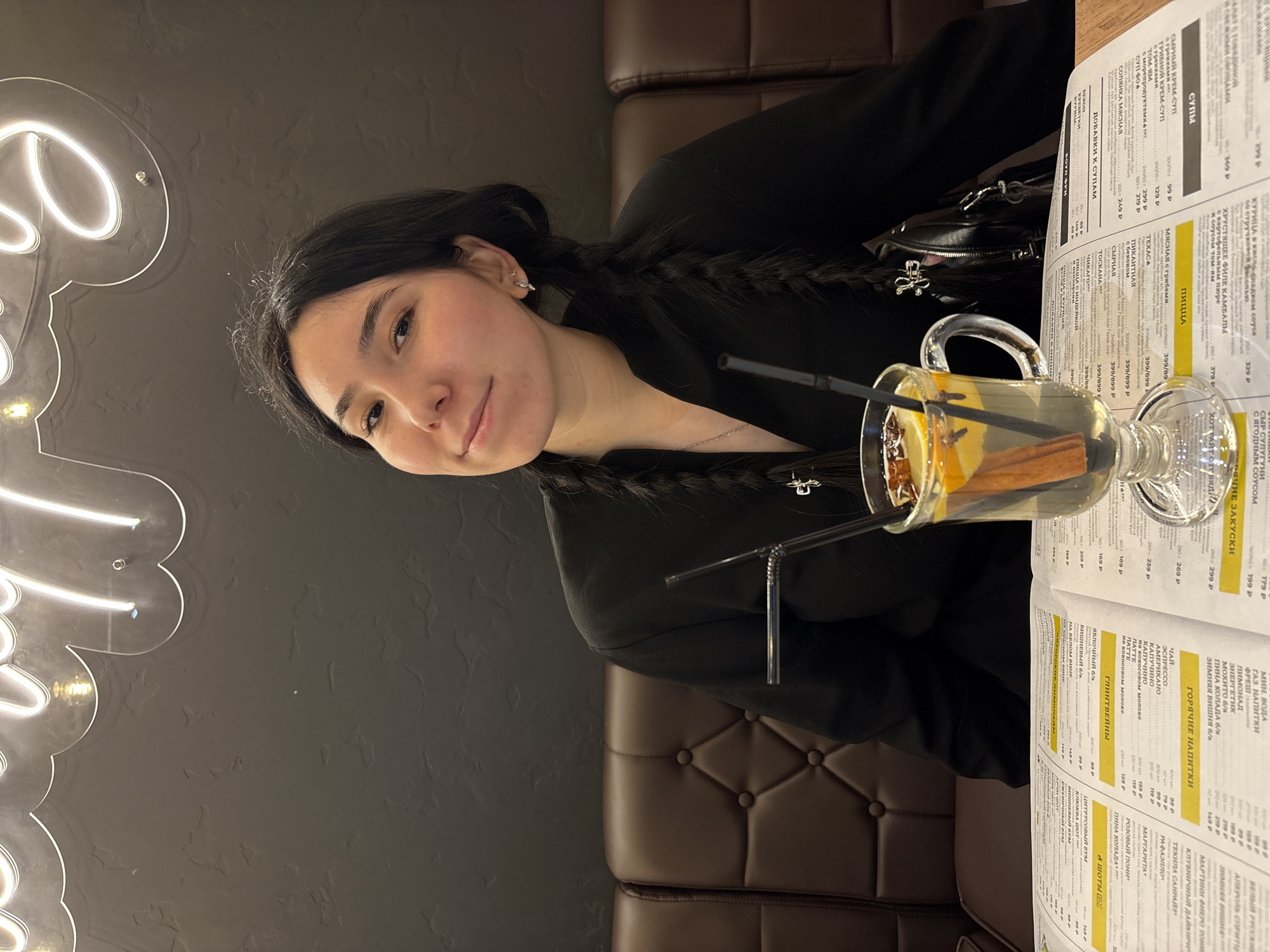 A young woman with dark hair, styled in braids, is seated at a table in what appears to be a restaurant or cafe, looking towards the camera with a subtle smile. She is wearing a black top or jacket and has small earrings.
On the table to her right, a clear glass mug contains a hot beverage, garnished with slices of orange or lemon, a cinnamon stick, and what appear to be star anise or cloves. A black straw is also in the mug. Adjacent to the drink, an open menu with extensive Russian text is prominently displayed. The menu features sections such as "СУПЫ" (Soups), "ПИЦЦА" (Pizza), "ГОРЯЧИЕ НАПИТКИ" (Hot Drinks), and "ГЛИНТВЕЙНЫ" (Mulled Wines). Specific items like "СЫРНЫЙ КРЕМ-СУП" (Cream Cheese Soup), "МЯСНАЯ с грибами" (Meat with mushrooms - under Pizza), and "КАПУЧИНО" (Cappuccino) are visible, with prices in Russian Rubles.
The background consists of a dark, textured wall. A glowing white neon sign with decorative, flowing script, partially visible on the left, adds to the ambiance. The woman is seated on a dark brown, tufted booth or bench. The lighting suggests an indoor, evening or night setting. The scene depicts a relaxed moment, possibly enjoying a meal or drinks.
