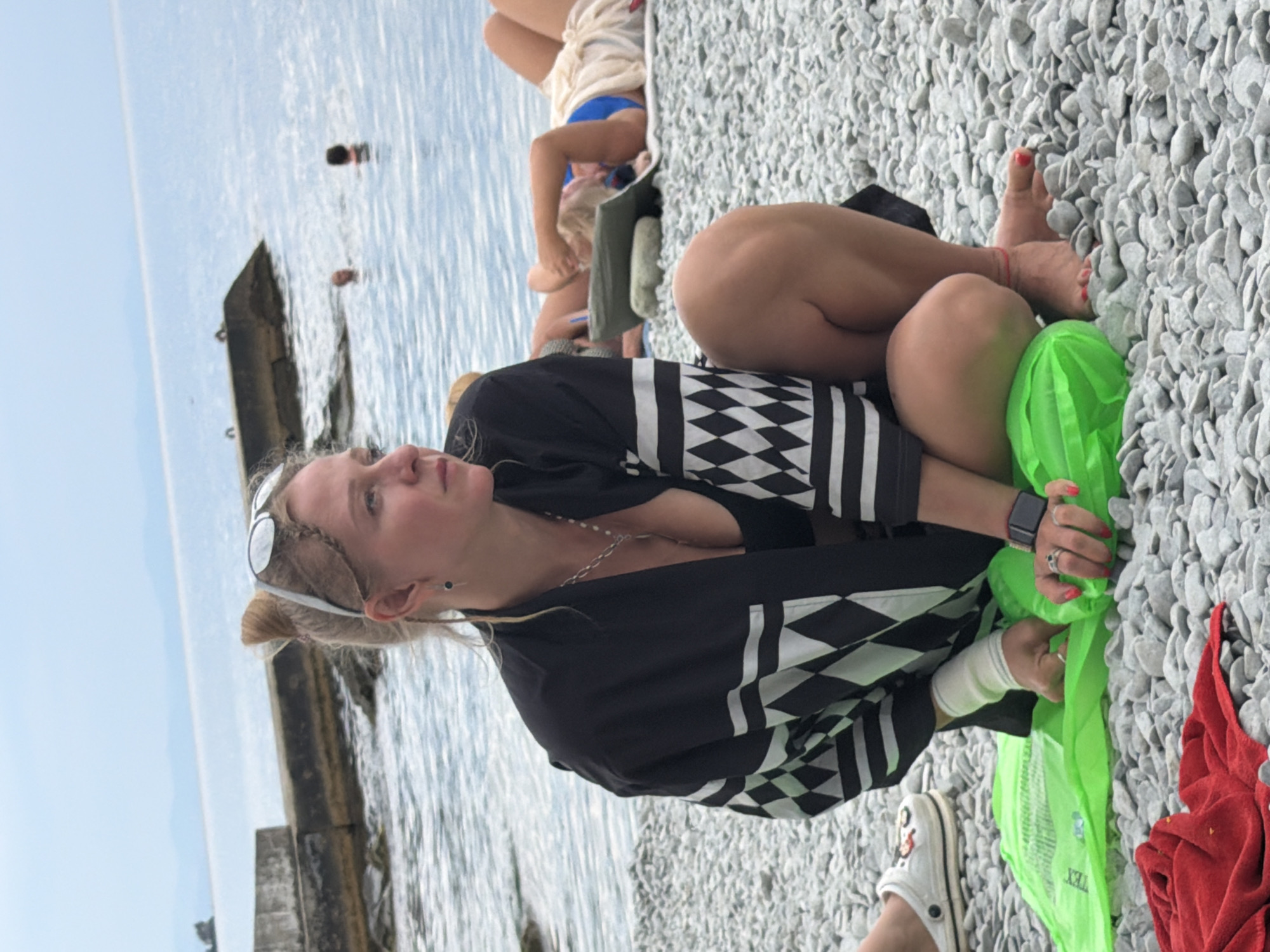 A woman with blonde hair tied up in a bun and sunglasses on her head sits on a pebble beach in Partenit, Ukraine. She is wearing a black and white patterned robe over a black bikini top. Her left arm is bandaged. She is holding a bright green inflatable object and has rings on her fingers, with red nail polish. To her left, a white Croc shoe with a design is visible. In the background, the sea is calm with a few people swimming. A concrete structure juts into the water from the left side of the frame. Another person is lying on the beach behind the main subject. A red towel is on the ground in the foreground.