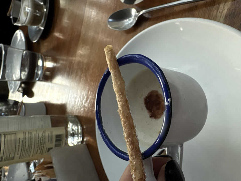 A white mug with a blue rim sits on a white saucer, holding a beige liquid with a dark swirl inside. A long, thin, cylindrical snack covered in granules is partially submerged in the liquid. A hand with black nail polish holds the snack. A spoon and a glass with a label are visible in the background on a wooden surface.