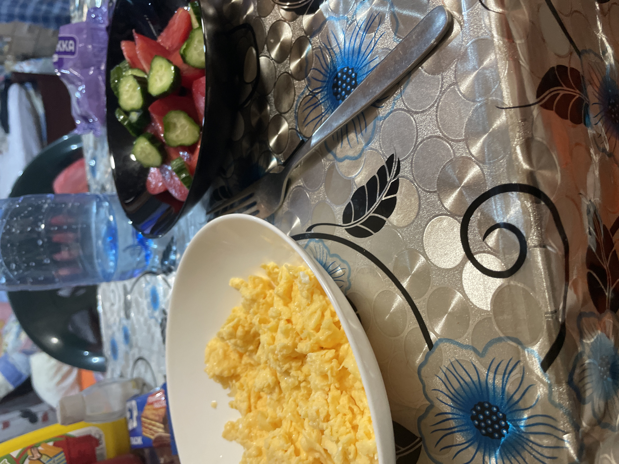 This media file captures a close-up, angled view of a meal set on a patterned tablecloth in what appears to be an indoor setting in Ihtiman, Bulgaria.
The main subjects are food items: a white, oval-shaped bowl in the foreground holds a large portion of fluffy yellow scrambled eggs. To the upper left, a black bowl contains a fresh salad consisting of sliced red tomatoes and green cucumbers. A silver-colored fork and knife are placed diagonally on the tablecloth, situated between the two bowls.
The table is covered with a distinctive patterned tablecloth. Its design features large blue flowers with black centers and delicate black lines extending outwards, along with swirling black leaf motifs. The background of the tablecloth is adorned with numerous overlapping silver-grey circles, giving a slightly metallic or brushed effect.
In the background, several other objects are partially visible. A clear plastic water bottle is present, as is a purple plastic item, possibly a bottle or packaging, which has the partial text "KKA" printed on it. Another blue and white package with the letters "CK" visible is also seen behind the scrambled eggs.
The activity depicted is the serving or preparation of a meal, likely breakfast or a light lunch. The lighting suggests an indoor environment, possibly artificial, creating some reflections on the tablecloth. The scene evokes a casual, home dining atmosphere.
