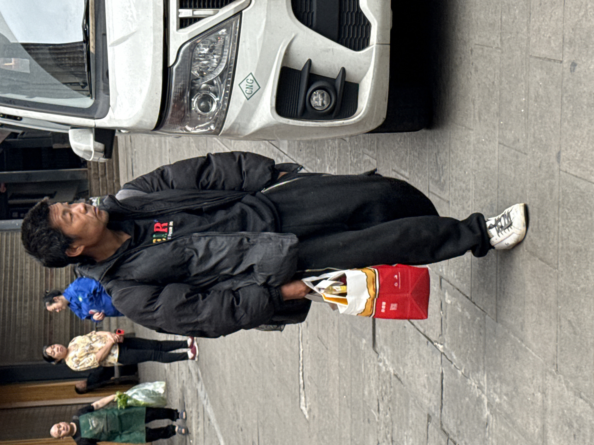 A man in a black jacket and pants walks on a grey stone-tiled sidewalk in Yuzhong District, China. He carries a red and white shopping bag in his right hand. A white vehicle is parked on the sidewalk near him. Behind him, three people walk along the sidewalk: a woman in a blue jacket, a woman in a patterned shirt and black pants, and another person in a green apron carrying a bag of produce.
