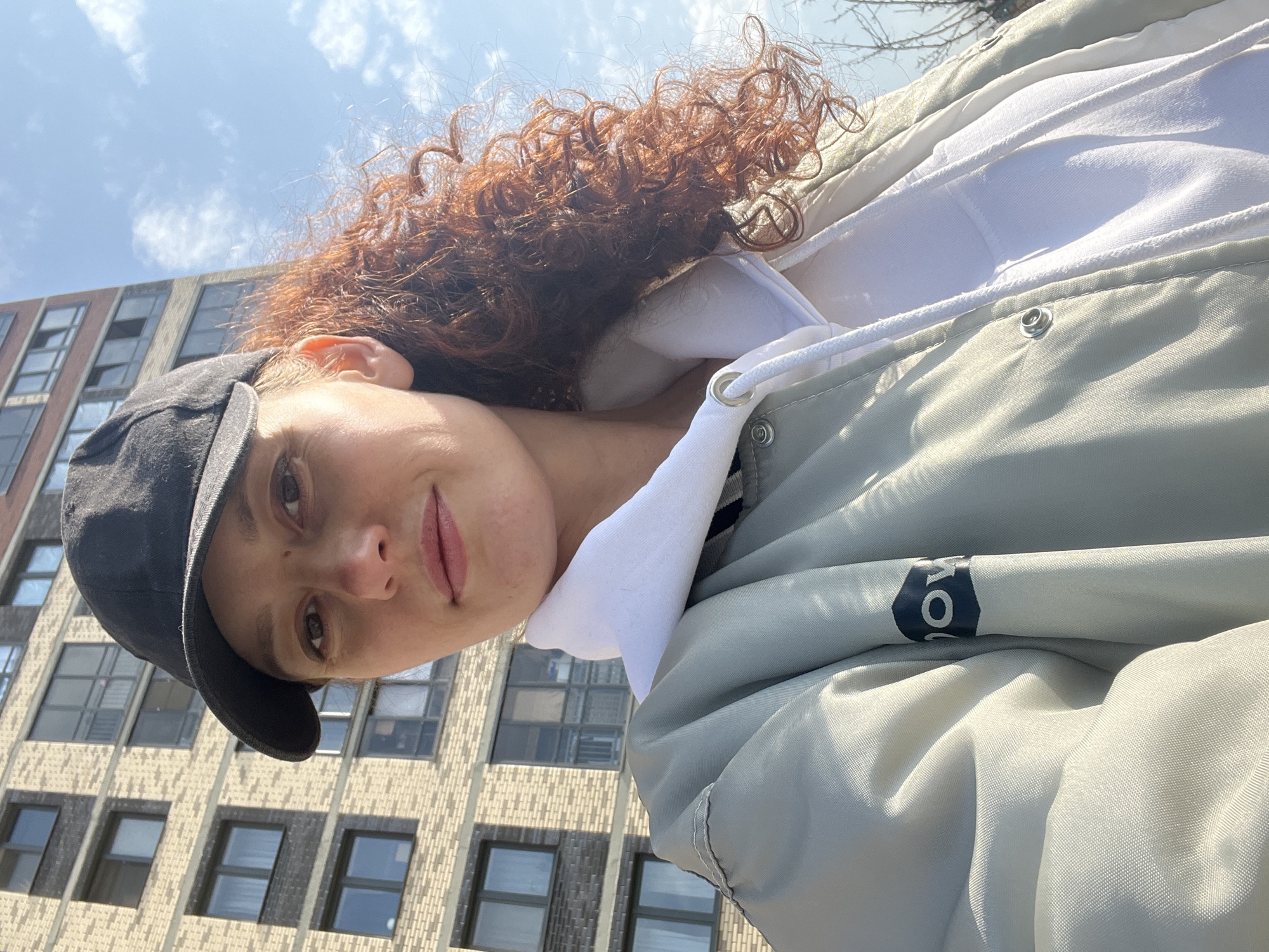 A person with curly red hair, wearing a black baseball cap, white hoodie, and a light grey jacket, is looking at the camera. The person is outdoors, standing in front of a tall building with many windows. The building appears to be in Saint Petersburg, Russia, based on architectural style, but this is not certain. The sky is blue with scattered clouds. The jacket has a buttoned collar and a logo on the shoulder.