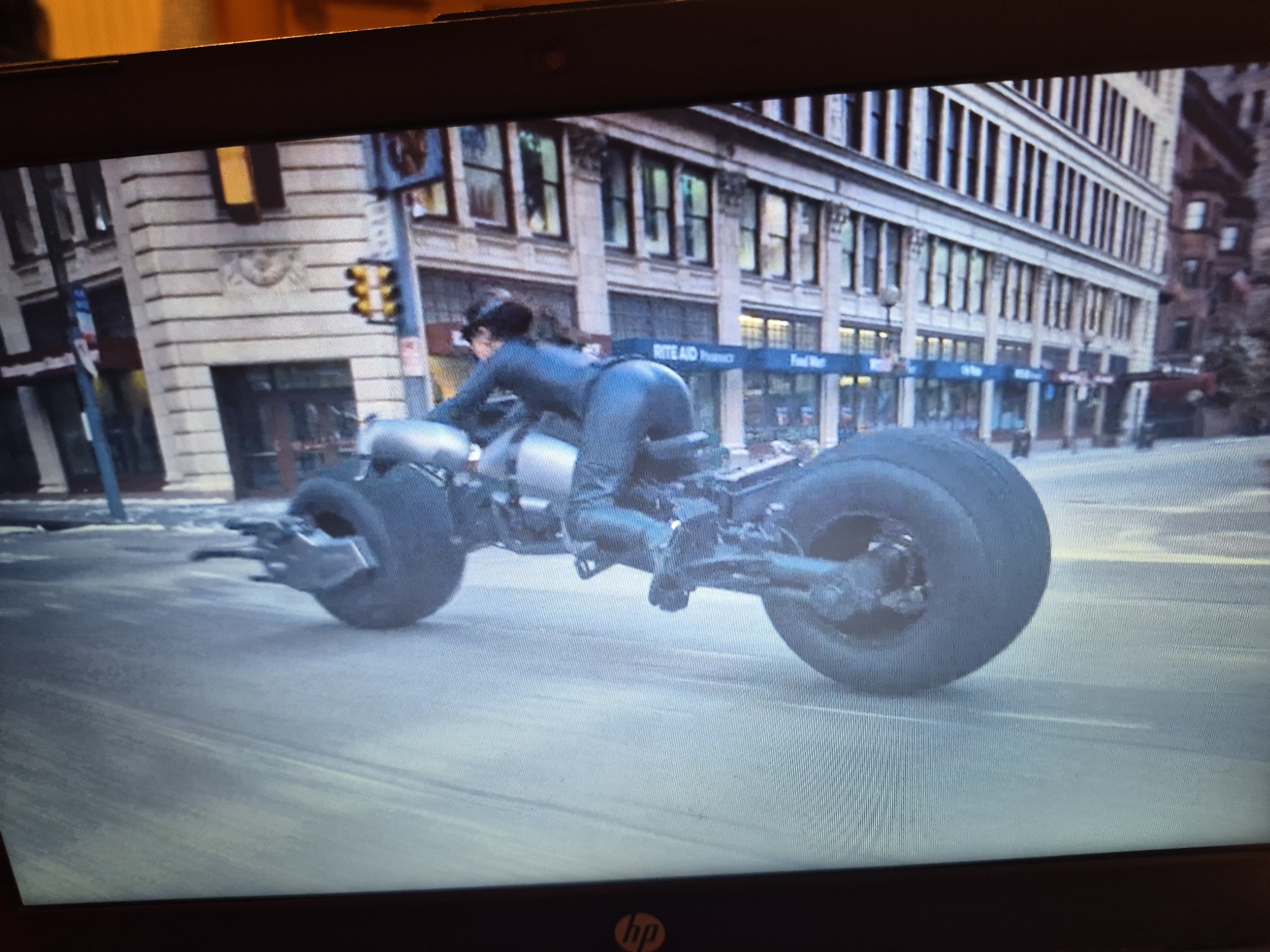 A person wearing a dark, form-fitting suit is riding a futuristic motorcycle in a city. The motorcycle has oversized wheels and appears to be a special-purpose vehicle. The rider is leaning forward, suggesting they are in motion. The background shows a multi-story building with a series of windows and architectural details. There is a traffic light visible on the side of the building, with its lights illuminated. The overall scene suggests an urban environment with the motorcycle in the foreground as the primary subject. The image does not provide enough information to determine the location as Sandwell, United Kingdom.
