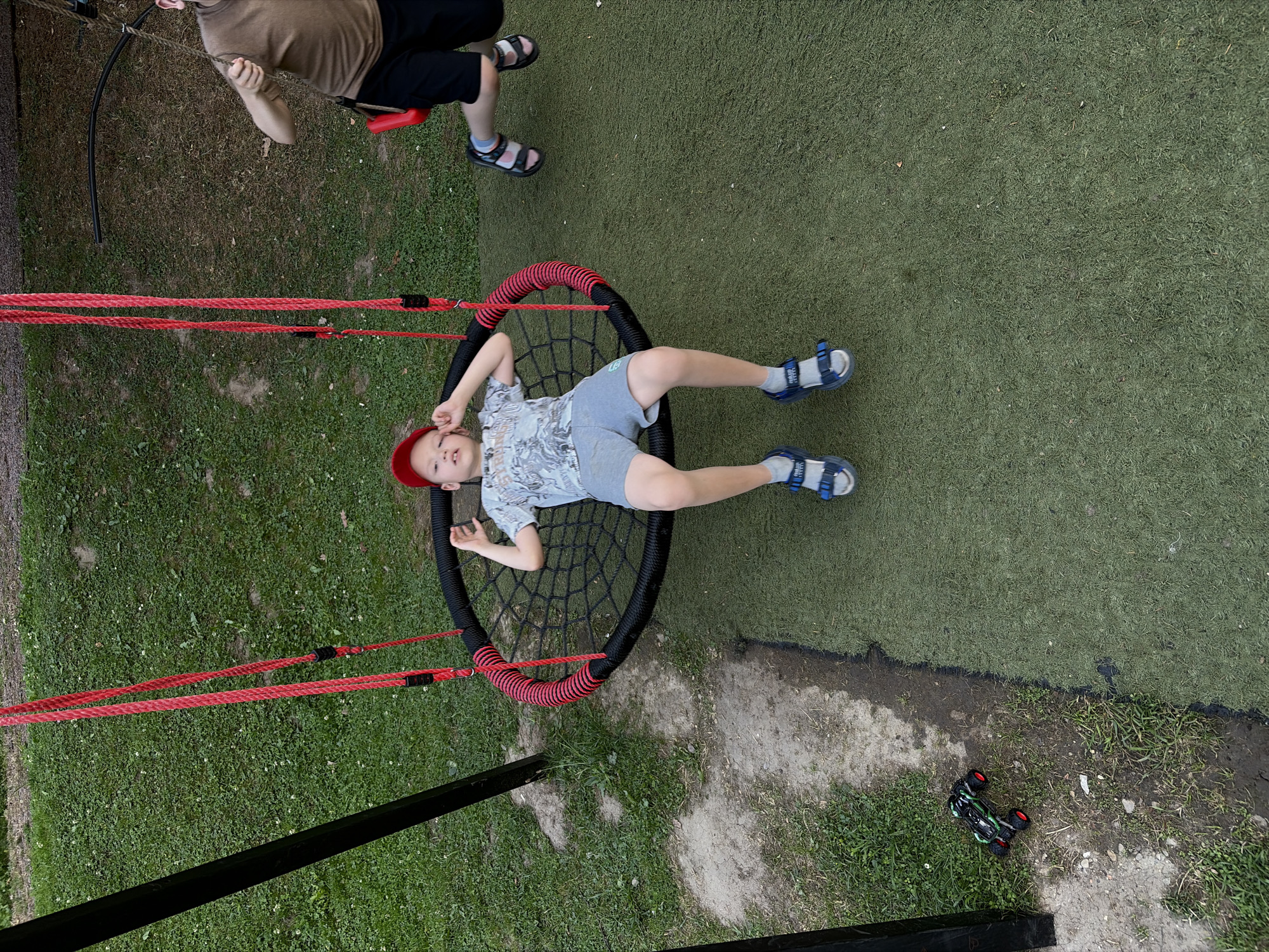 A medium shot captures two young boys on a playground. The boy in the foreground, wearing a red cap, a light-colored t-shirt with a pattern, and grey shorts, is seated on a circular black net swing with red rope suspension. He is looking slightly to his left, with his arms resting on the sides of the swing. His legs are bent at the knees, and he is wearing grey socks and blue sandals.
To the right of the foreground boy, another boy is visible on a red swing. He is wearing a brown t-shirt and black shorts, and his legs are extended as he swings. His hands are holding the swing chains.
The playground has a surface of artificial turf in a vibrant green color. To the left of the foreground boy, a small black and green toy remote-control car with red wheels rests on a patch of dirt and grass. In the background, a mix of grass and bare earth is visible. The overall scene suggests a recreational outdoor setting, likely a park or a backyard playground.
