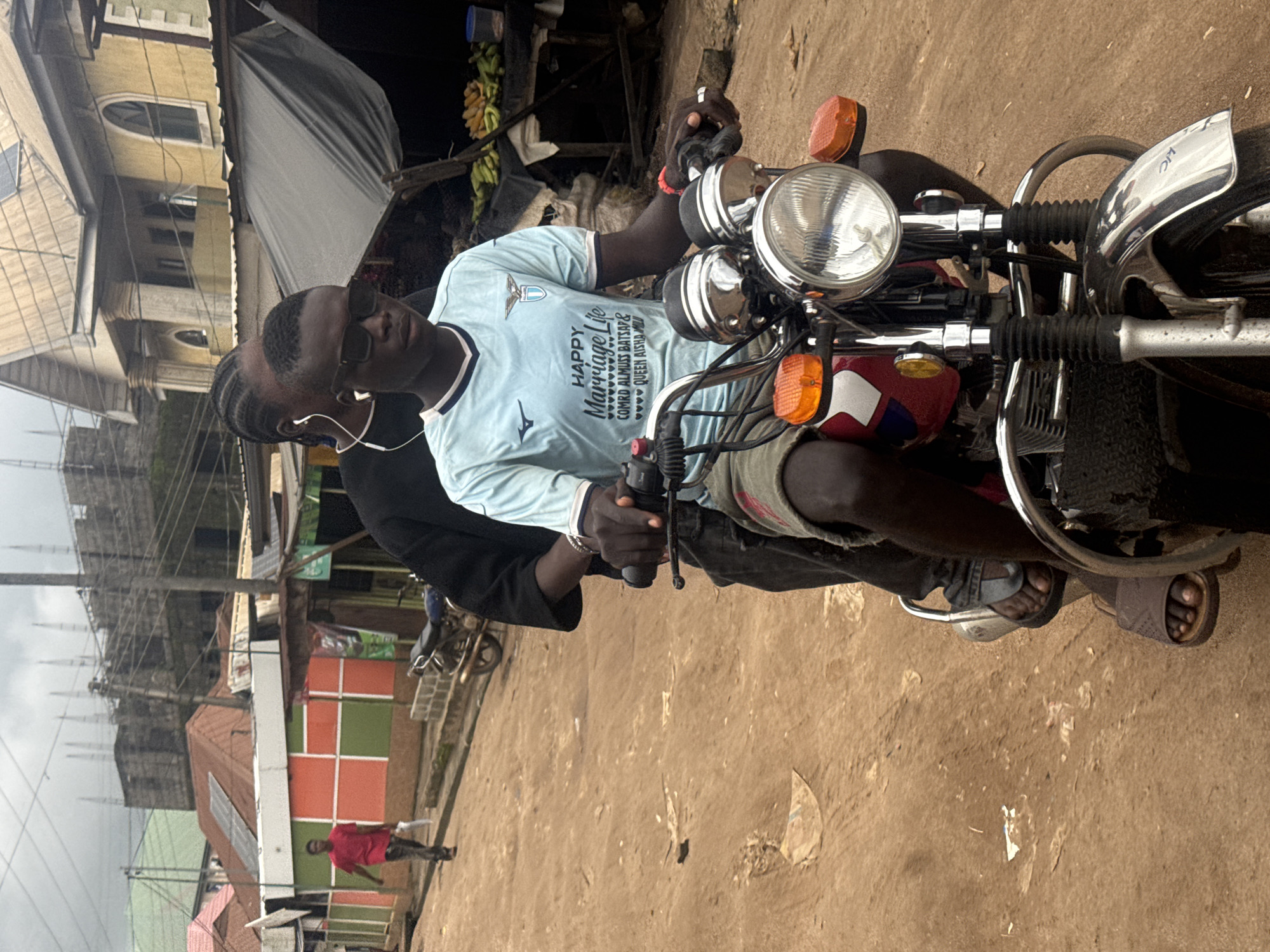 In Ihiagwa, Nigeria, two individuals are riding on a motorcycle down a dirt road. The person in front, wearing sunglasses and a light blue t-shirt with "HAPPY Marriage Life" printed on it, is driving the motorcycle. The second person, whose face is not fully visible, is seated behind the driver. They are both dressed in casual clothing and appear to be engaged in a conversation. The environment is a dusty street with buildings on either side. In the background, there are more buildings under construction, some with visible scaffolding, suggesting ongoing development. The sky is overcast, indicating a cloudy day. The scene depicts a common mode of transportation in the region.