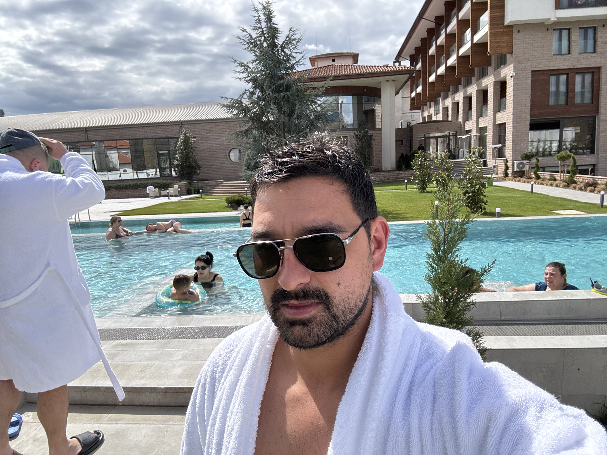 The photo depicts a man in a white robe and sunglasses taking a selfie at what appears to be a resort pool in Panagyurishte, Bulgaria. Behind him, people are swimming and relaxing in the pool. A building with a modern design is visible in the background, and trees and well-maintained landscaping surround the pool area. The weather seems bright and partly cloudy. Another man wearing a bathrobe and a cap is walking by on the left.