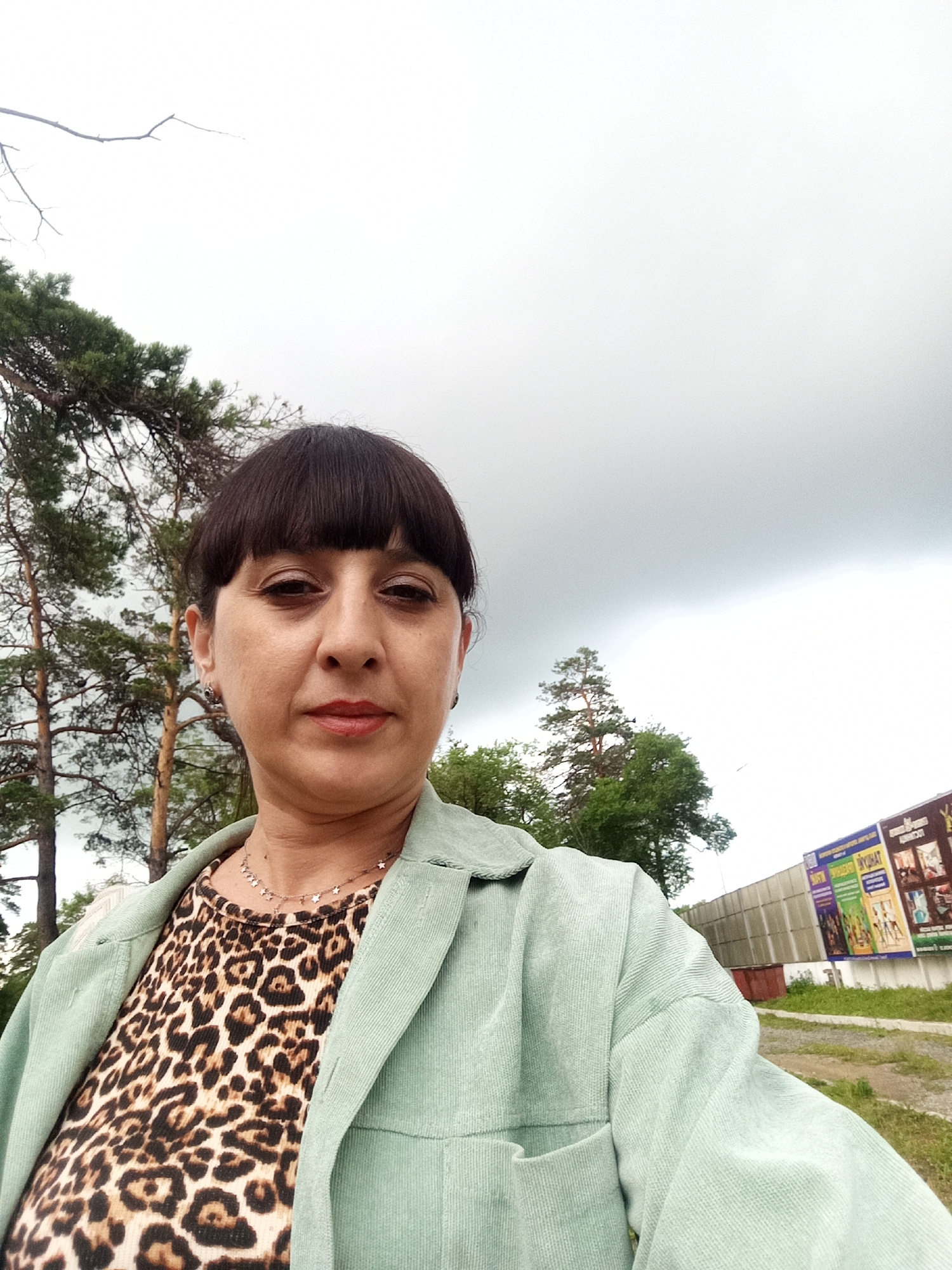 A woman is taking a selfie outdoors. She has dark hair with bangs and is wearing a leopard print shirt and a light green jacket. In the background, there are trees, a cloudy sky, and a billboard with advertisements. The scene appears to be a public area, possibly a park or a street in Belogorsk, Russia, given the billboard's presence.