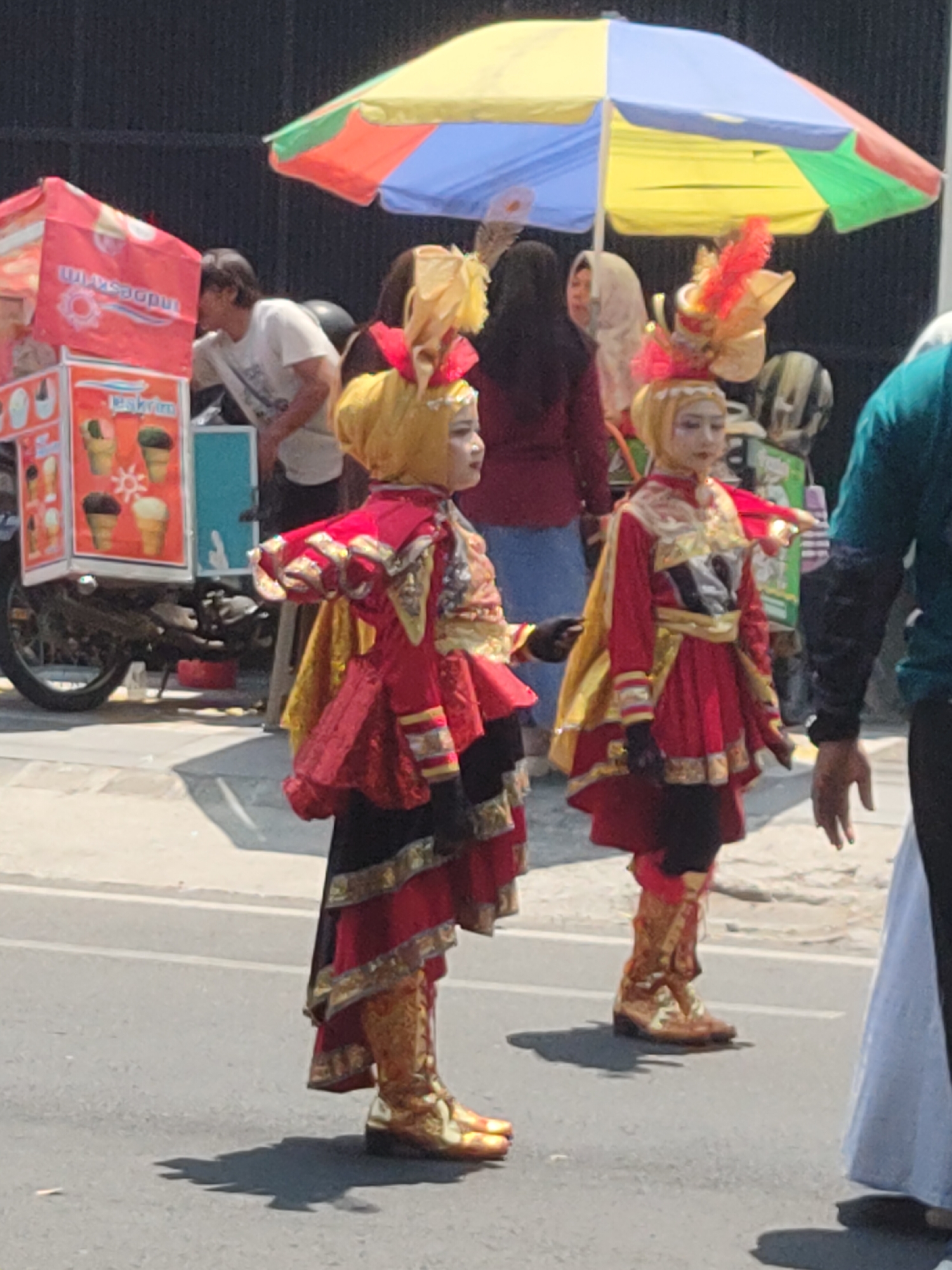 In Ngasem, Indonesia, two young girls in elaborate red and gold traditional costumes stand on an asphalt road. The girl on the left wears a yellow headdress with red and gold accents, a red tunic with gold trim, and black leggings with red and gold patterned boots. The girl on the right has a similar costume, with a yellow headdress adorned with a prominent red feather. Behind them, an ice cream cart with a colorful umbrella shades several people. One person, wearing a white t-shirt, is partially visible, leaning towards the cart. The environment appears to be outdoors, possibly during a daytime event or festival.