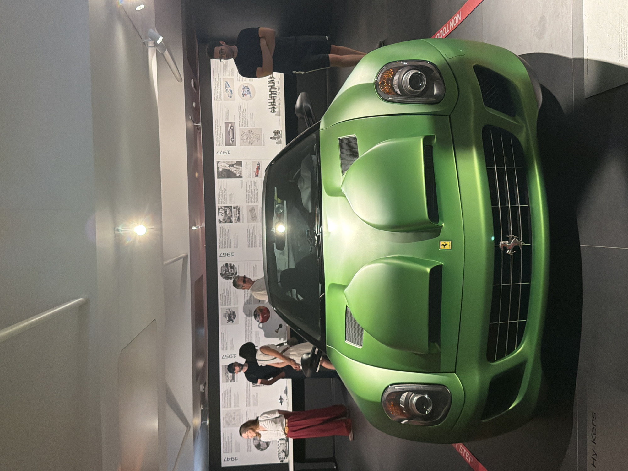 The image shows the interior of a museum exhibit, likely the Ferrari Museum in Maranello, Italy. In the foreground, a bright green Ferrari is prominently displayed, viewed from the front. The car has a distinctive design with large air intakes on the hood. Behind the car, several people are observing the exhibit. A timeline display with various dates and images is mounted on the wall behind them. To the right, a man wearing a black t-shirt and shorts stands with his arms crossed. The lighting suggests an indoor setting with spotlights illuminating the exhibit. The floor is dark, and red tape barriers are visible on the floor in front of the car.