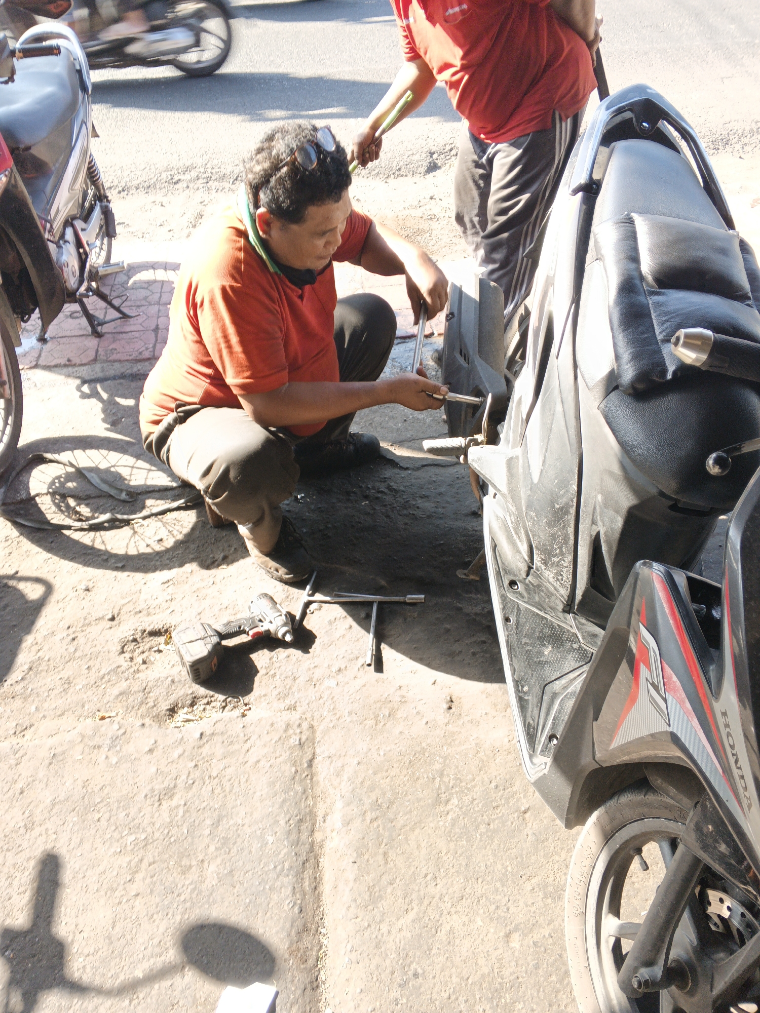 In Sukawati, Indonesia, a man in an orange t-shirt and black pants is squatting on the ground next to a parked motorcycle, appearing to be repairing it. He is holding a tool, likely a wrench or socket, and is actively working on the motorcycle's rear wheel area. Another individual, wearing a red t-shirt and grey and black striped pants, stands behind the first man, holding a tool that resembles a broom handle or a stick, possibly assisting or observing. The scene is outdoors, with a dusty, sandy ground surface and visible shadows cast by the sun. Other motorcycles are partially visible in the background, suggesting a roadside repair shop or a similar service area. The overall environment is consistent with a warm, sunny day in a bustling urban or semi-urban setting.