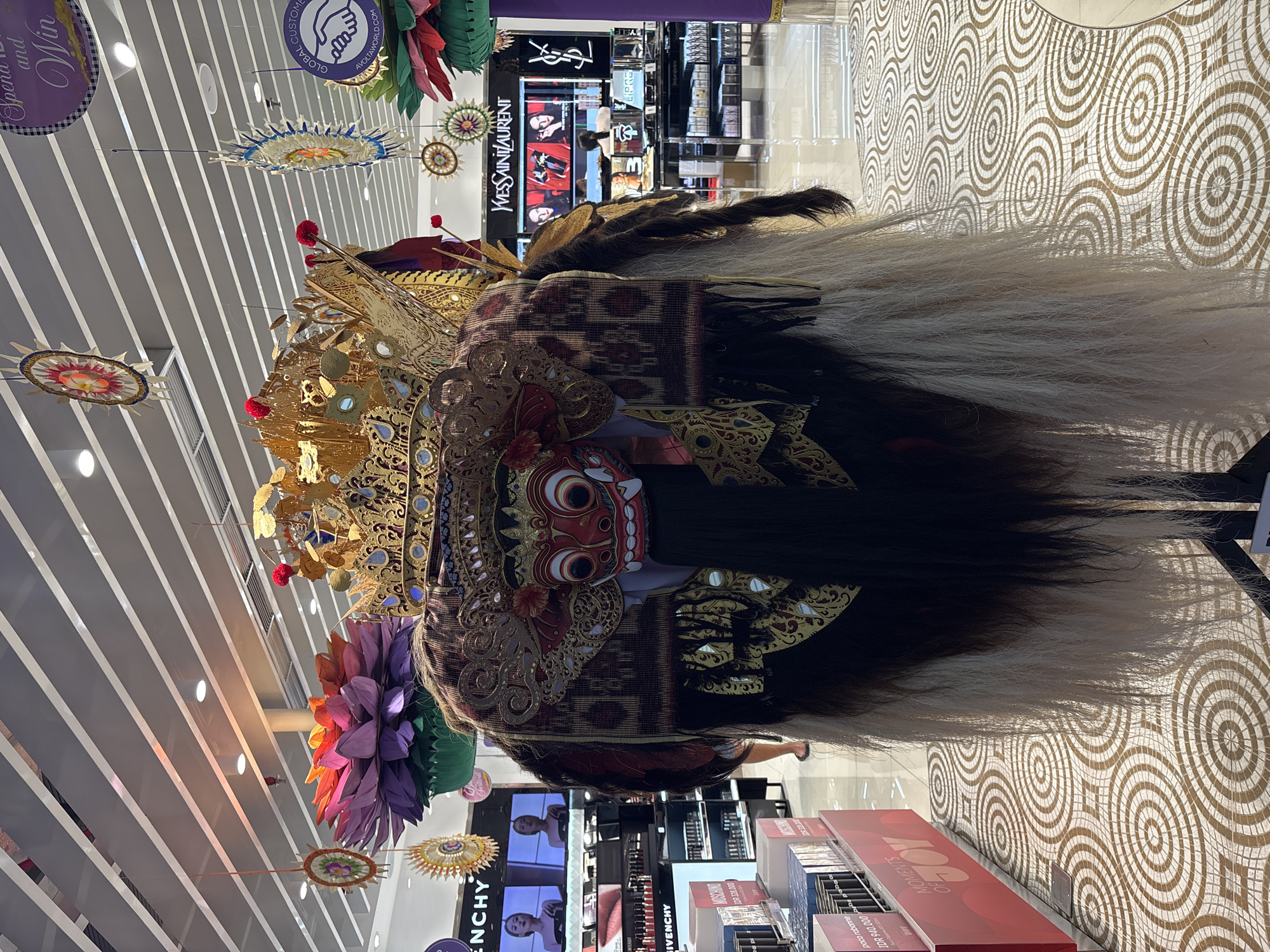 The image captures a cultural display inside a commercial area, specifically within a store featuring brands like Yves Saint Laurent, Givenchy, and Paco Rabanne. The focus of the display is a traditional Balinese Barong mask, characterized by its ornate golden crown, red and gold face, and long white and black hair. Colorful paper decorations hang from the ceiling, which has white horizontal stripes. The floor has a patterned, concentric circle design. The location appears to be in Kuta, Indonesia, given the Balinese cultural element.