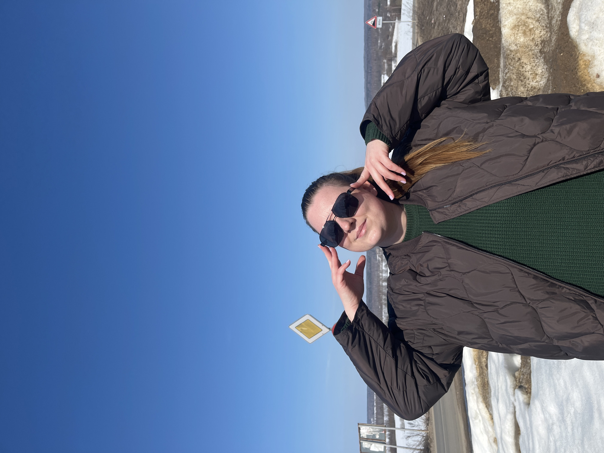 A female subject stands centrally in a mid-shot outdoor scene in the city of Zubovo, Russia, under a clear, cloudless blue sky. She wears dark sunglasses, a dark green knitted sweater, and a dark brown quilted bomber jacket. Her long, light brown hair is visible on her right shoulder. Her hands are raised to the sides of her head, with fingertips positioned near her temples, and she looks slightly upward and to her left with a subtle smile. To the left of the subject, a white diamond-shaped road sign with a yellow interior, indicating a priority road, is visible on a post. Further in the background on the right, a triangular warning sign with a red border and the text "800м" is present. The ground around the subject and extending into the mid-ground consists of patches of melting snow interspersed with bare brown earth, bordering a visible section of a paved road on the left. A distant tree line (forest) forms the horizon.