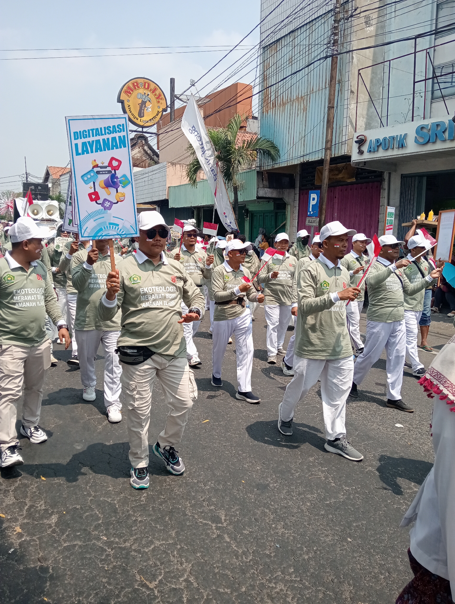 In Demak, Indonesia, a group of men wearing matching olive green long-sleeved shirts and white baseball caps are participating in a street parade. The shirts have a logo with green and white imagery and text that appears to be "EKOTEOLOGI MERAWAT BUMI AMANAH ILMU". One man in the foreground holds a sign that says "DIGITALISASI LAYANAN" and features various icons representing digital services. Many of the men are holding small Indonesian flags, and some have white face paint on their cheeks. They are walking on a paved street lined with buildings. One building in the background has signage that includes "APOTIK SRI". The sky is clear and blue. The overall scene depicts a civic or promotional event.
