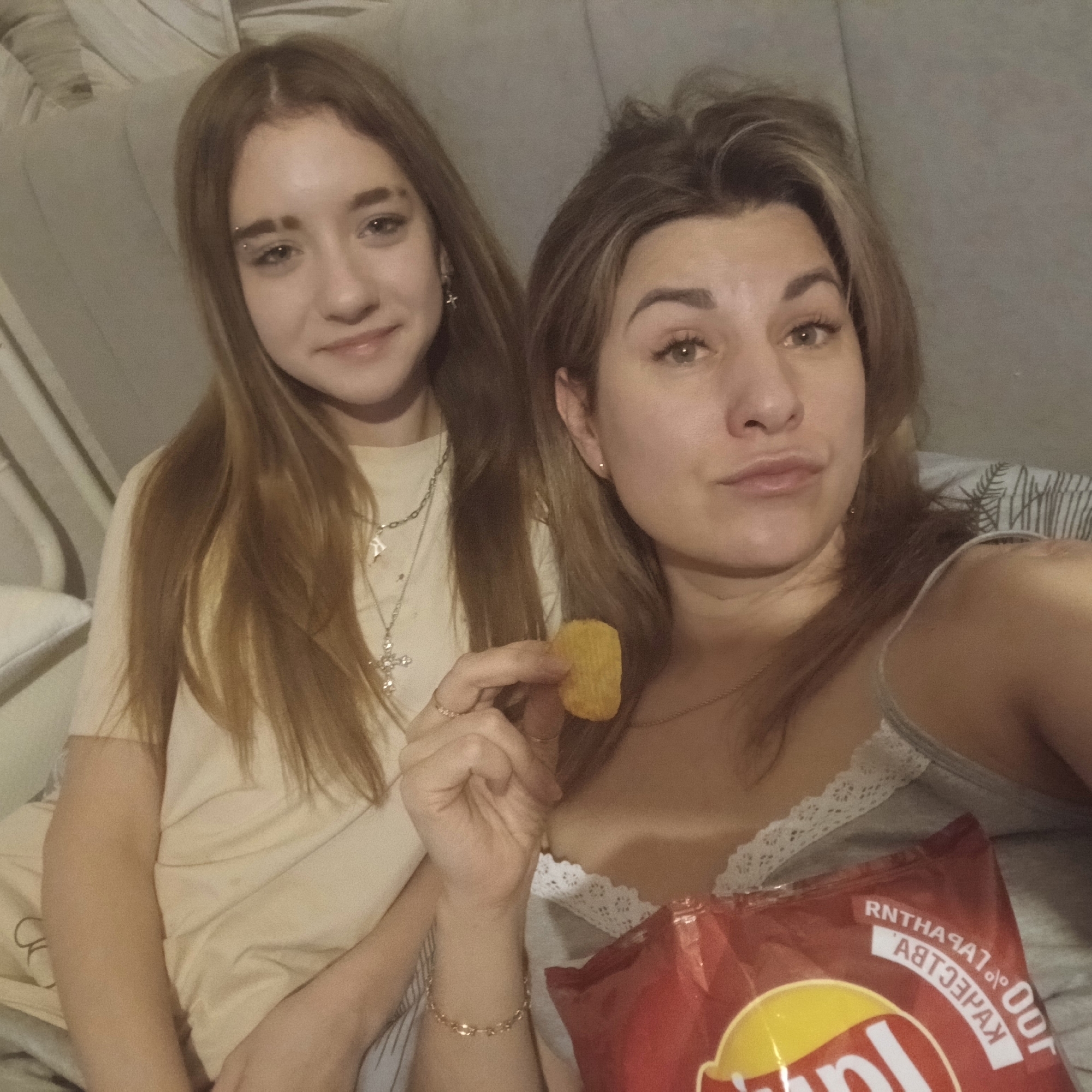 Two women are sitting or lying down on a gray fabric-covered surface, possibly a bed or couch. The younger woman is on the left side of the frame. She has long, straight brown hair, fair skin, and is wearing a light-colored t-shirt and a silver necklace with multiple pendants. The older woman is on the right side and is holding a potato chip between her fingers. She has brown hair with blonde highlights, fair skin, and is wearing a gray tank top with white lace trim. A red bag of Lay's potato chips is partially visible in front of her. The backdrop appears to be a plain, light-colored wall or headboard. The overall lighting is soft and diffused. The image was not necessarily taken in the city of Izhevsk, Russia. There is no information available that indicates that.