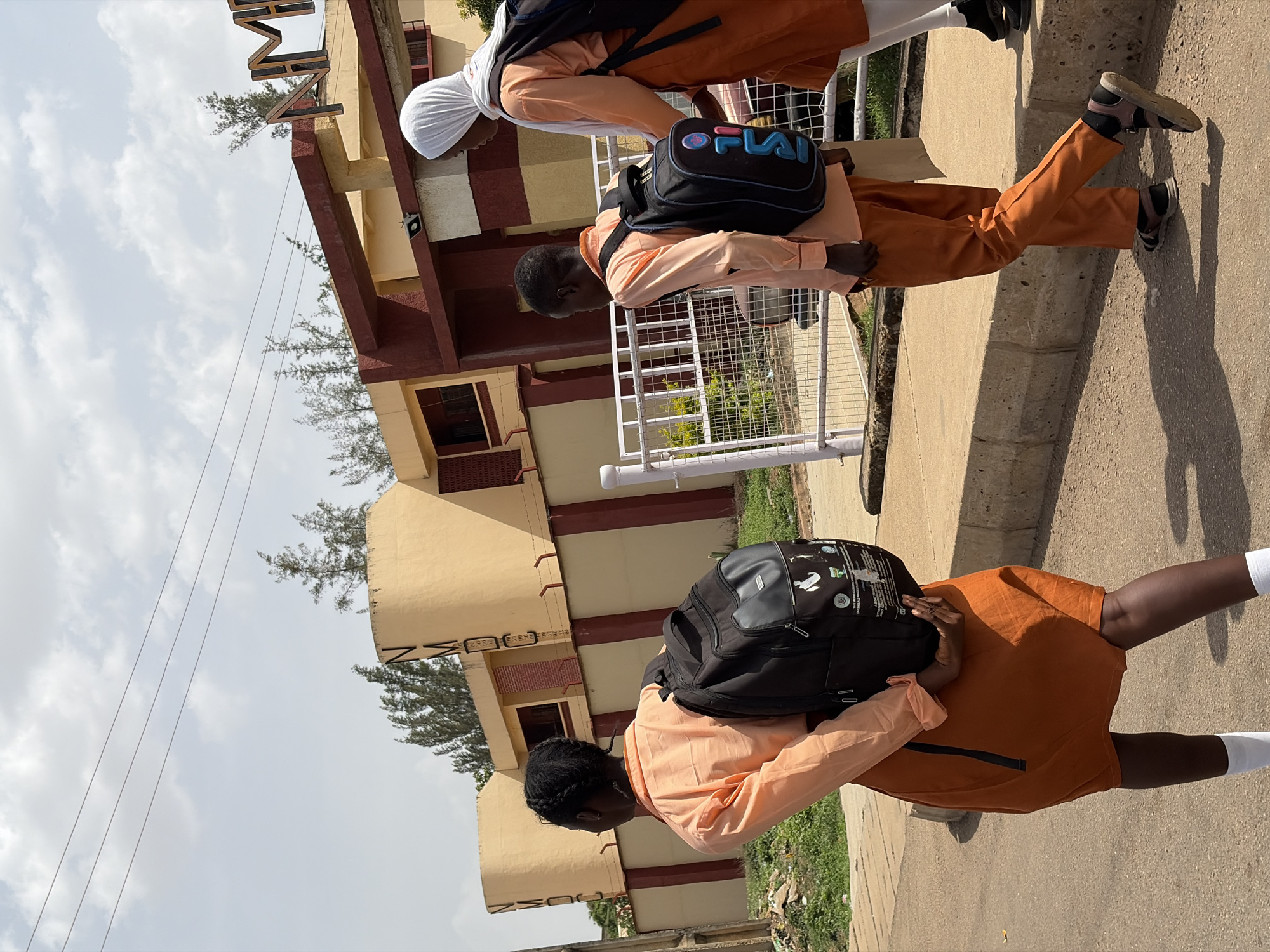 Three individuals, likely students, walk along a concrete path in Rafin Par, Nigeria, under a bright, partly cloudy sky. The scene depicts an exterior setting, possibly near a school or institutional building.
In the foreground, a person with braided hair is seen from behind, wearing a black backpack, a light peach long-sleeved shirt, and an orange skirt or dress, with white socks visible. Further down the path, two more individuals walk side-by-side. One person, positioned slightly ahead, wears orange trousers, a light peach long-sleeved shirt, and dark sandals, carrying a black backpack adorned with a prominent "FILA" logo in blue and pink letters. Next to them, a person in a white head covering (hijab), orange trousers, and a light peach long-sleeved shirt walks along. All three are carrying black backpacks, suggesting they might be commuting to or from an educational institution.
To their left, a metal fence separates the path from a green area with small plants. In the background, a multi-story building with a light yellow facade and reddish-brown columns or trim is visible. On the upper part of the building, the letters "AMIN" are clearly discernible from a sign. Another set of letters, "NSUC" or "NSCO," is partially visible on a lower section of the building. Scattered trees and shrubs are seen around the building and in the distance. Overhead electrical wires stretch across the sky. The bright daylight indicates it is likely midday or early afternoon, casting strong shadows on the ground.