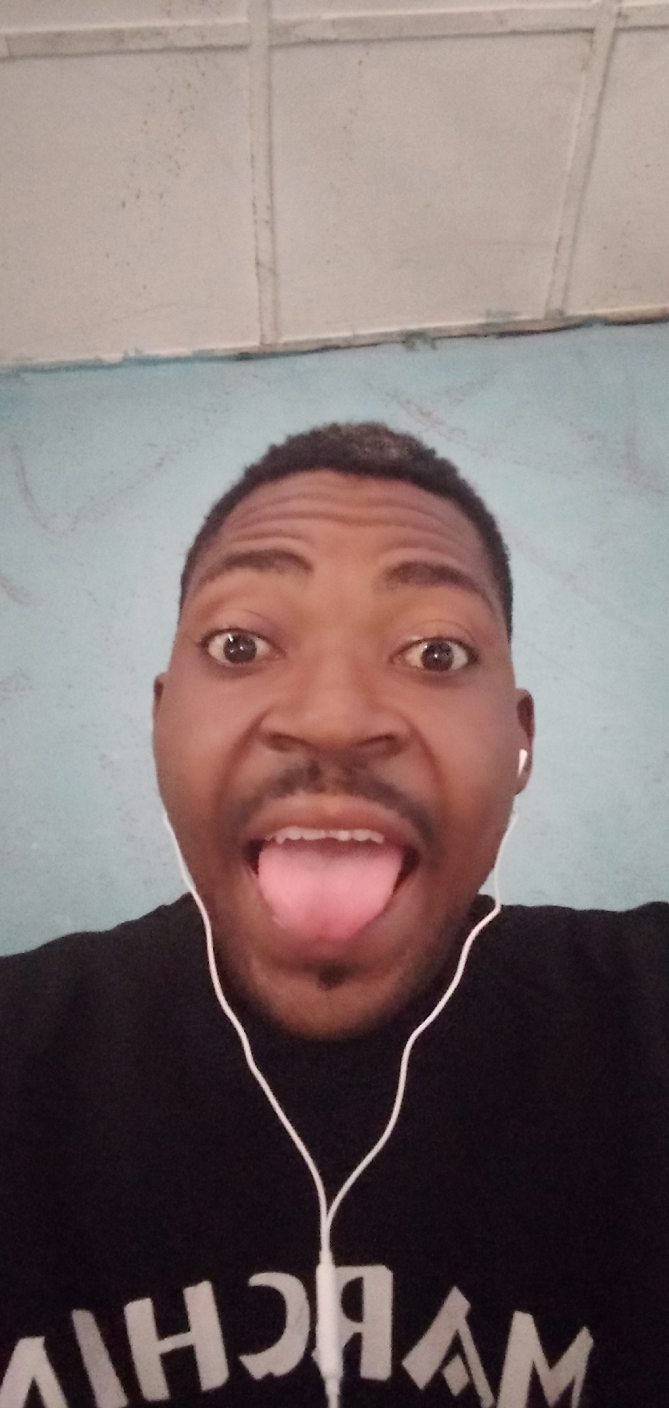 A man with dark skin, short hair, and a mustache is looking directly at the camera with his mouth open and tongue protruding. He is wearing a black shirt with white text on it and white earphones. The background consists of a light blue wall and a white ceiling with visible seams. The image was taken at Ejule, Nigeria.