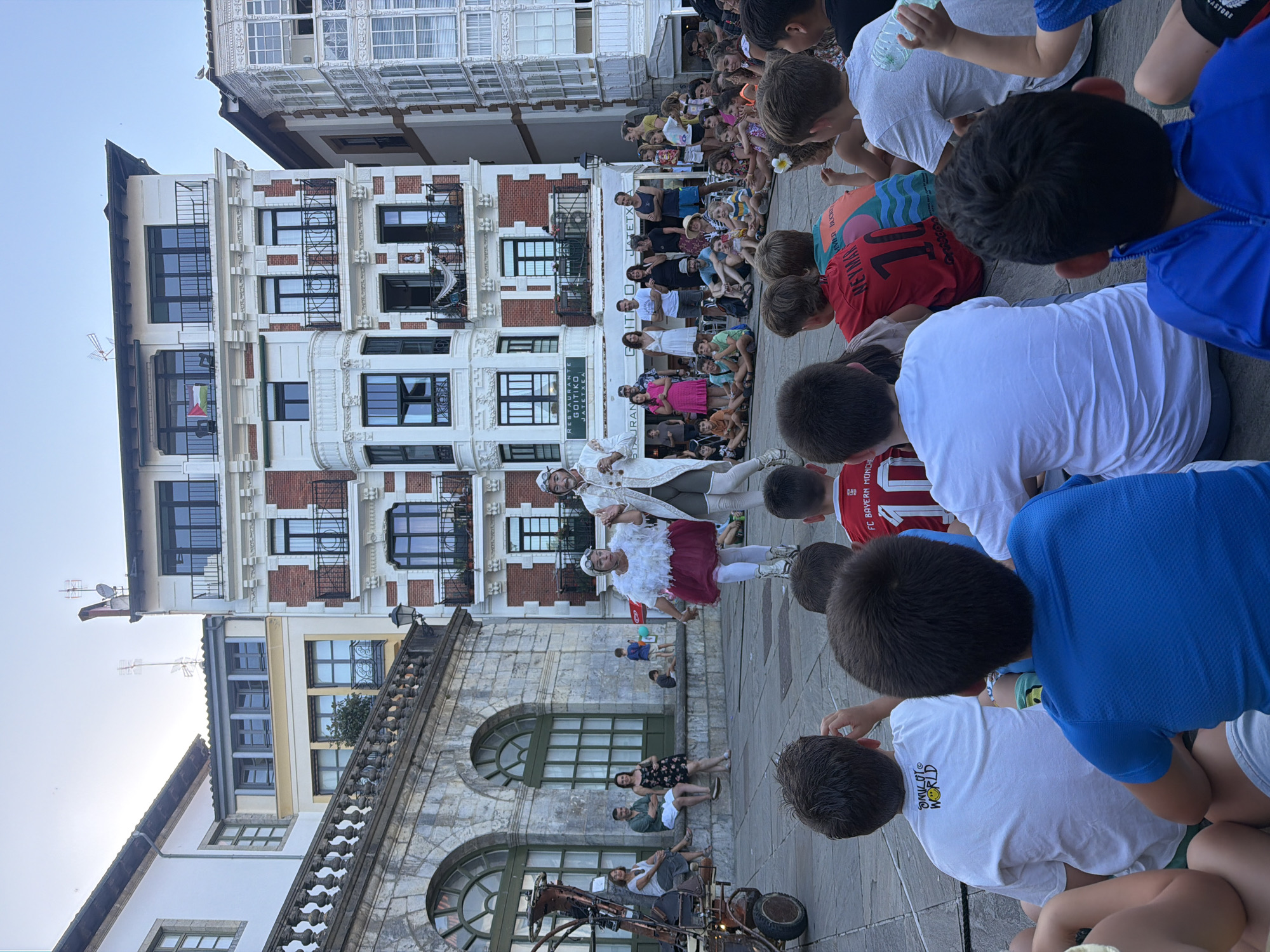In Lekeitio, Spain, a street performance is underway in a public square with a stone building on the left and ornate apartment buildings in the background. The focus is on two performers dressed in elaborate costumes, one in white with a feathered collar and the other in a white coat, white leggings, and goggles. They are performing for a crowd of onlookers, primarily children seated on the ground in the foreground, facing away from the camera. The children are wearing casual clothing, with some wearing soccer jerseys. Other spectators, adults and children, are visible further back, some standing and some seated on a stone ledge. A large, steampunk-like mechanical structure is partially visible on the far left. The atmosphere appears festive, with the performance drawing attention from the gathered audience.