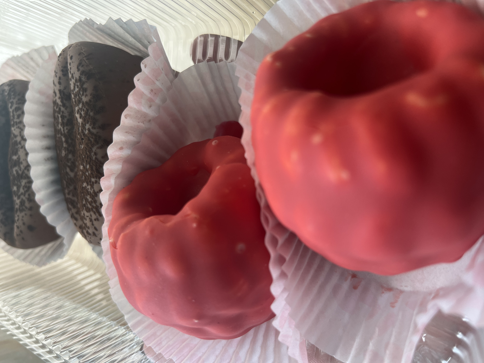 This image shows a close-up view of several pastries in a clear plastic container. The pastries are arranged in a row, each nestled in a fluted paper liner. In the foreground, two pastries are coated in a bright red icing. One is slightly out of focus in the lower right, and the other is more in focus in the center. Both red pastries have a somewhat irregular, folded shape. Behind the red pastries, two chocolate-covered pastries are visible, dusted with what appears to be cocoa powder or chocolate shavings. These chocolate pastries are also in paper liners. The background shows the textured, ridged interior of the plastic container and other similar containers stacked behind it. The lighting suggests the pastries are indoors, possibly in a bakery or food display. There are no people or explicit actions depicted. The specific location in Desyatikhatka, Russia, cannot be determined from the visual information provided in the image.