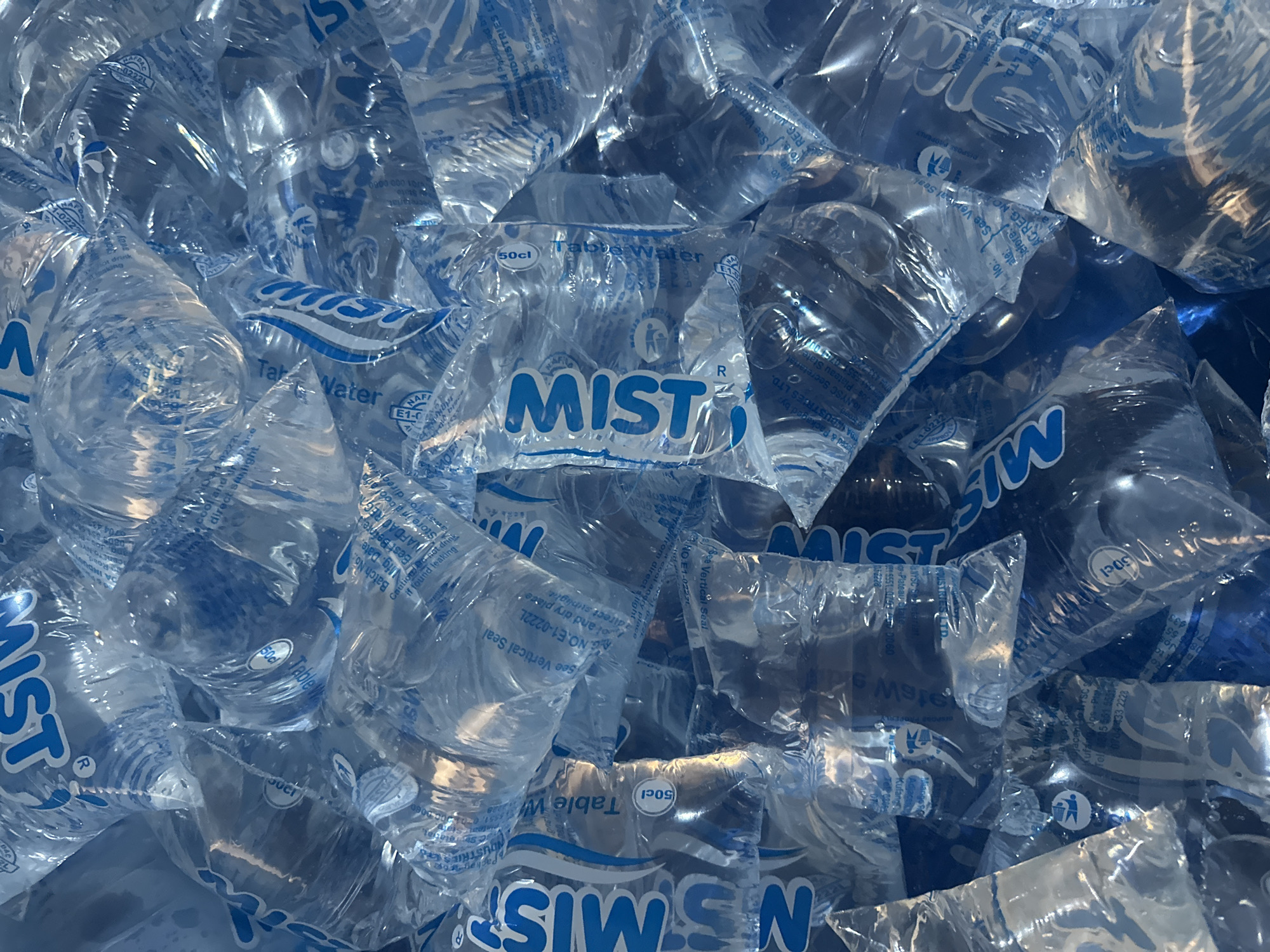 The image presents a close-up, eye-level view of a pile of "MIST" brand table water sachets. The sachets are transparent, filled with water, and feature a blue and white logo. The logo prominently displays the word "MIST" in a stylized font. Other text on the sachets includes information such as "Table Water," "50cl," batch number, manufacturing date, and best before date.
The text also identifies the manufacturer as MIDA INDUSTRIES LTD., located at No. 10 NYSC Secretariat, Adjacent to NYSC in Kufang, Jos-Plateau State, Nigeria. Information about disposal and precautions (storing in a cool, dry place away from direct sunlight; not drinking if found leaking) is visible. The sachets are stacked haphazardly, with some overlapping and obscuring others.