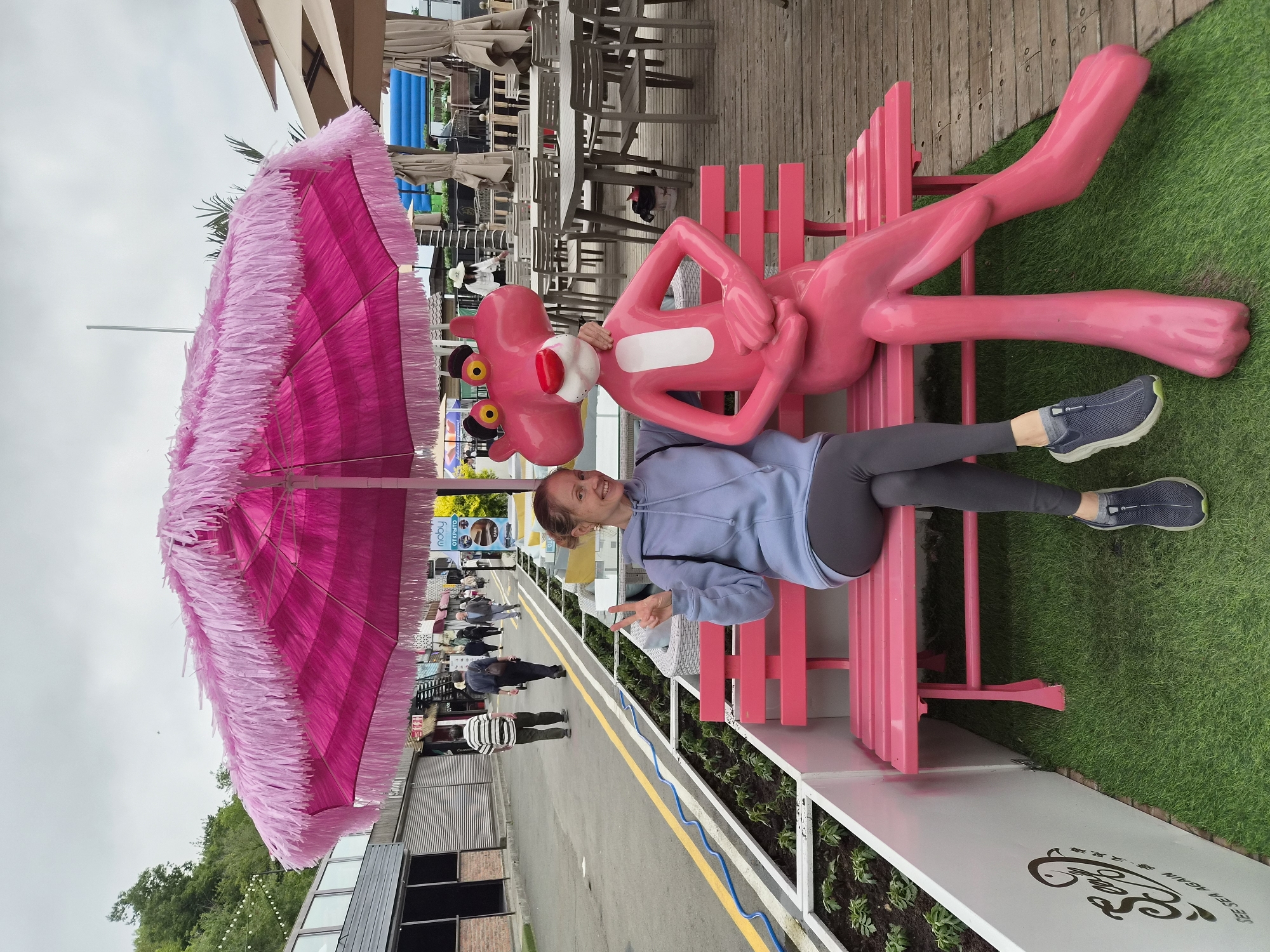 A woman with blonde hair pulled back, wearing a light blue hooded sweatshirt and dark grey leggings, sits on a pink park bench next to a life-sized pink panther statue. She is smiling and has her left hand raised with two fingers extended in a peace sign. The pink panther statue is seated, with its left arm resting on the woman's shoulder. A large, bright pink umbrella with fringed edges is propped up behind them. The bench is situated on an area of artificial grass, with a white planter box containing small green plants to the left. Behind the bench, a wooden deck with tables and chairs is visible, with a street and buildings in the background. The sky is overcast. The scene appears to be outdoors in a public area, possibly a promenade or a seating area outside a cafe or restaurant. The text "SEE SEA AGAIN" is visible on the planter box to the left. This location is in Vladivostok, Russia.