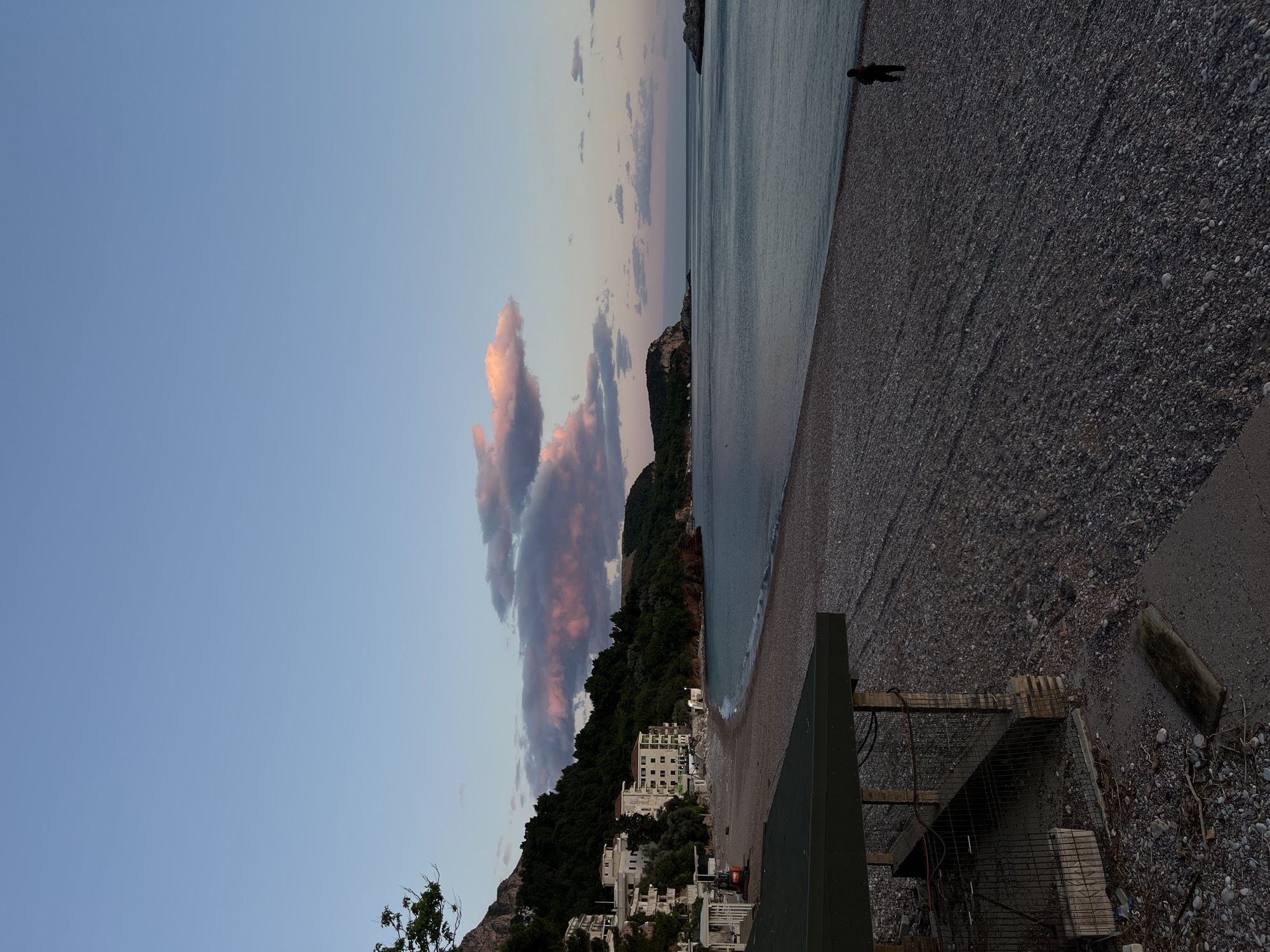 This media file depicts a tranquil coastal scene in Sveti Stefan, Montenegro, captured during dusk or dawn. The sky, a clear blue at the top, gradually transitions into soft pink and orange hues towards the horizon, punctuated by elongated, colored clouds.
The calm sea occupies the right side of the image, its surface gently rippling and reflecting the muted colors of the sky. A long, pebbled beach stretches along the coastline, with dark gray and brown stones making up most of its texture. A single individual is visible as a dark silhouette standing near the water's edge on the beach, facing the sea, appearing to be observing the view or walking.
On the left, a densely forested hill, rich in dark green foliage, slopes down to meet the shoreline. Nestled at the base of this hill are several white, multi-story buildings with reddish-brown roofs, characteristic of Mediterranean architecture. In the bottom right foreground, there are some structures, including what appears to be a dark green barrier or wall, and part of a wooden fence or frame, alongside more pebbles and some scattered debris on the beach. The soft, diffused light and warm cloud colors suggest the setting sun, imbuing the scene with a peaceful and serene atmosphere. No visible text is present in the image.