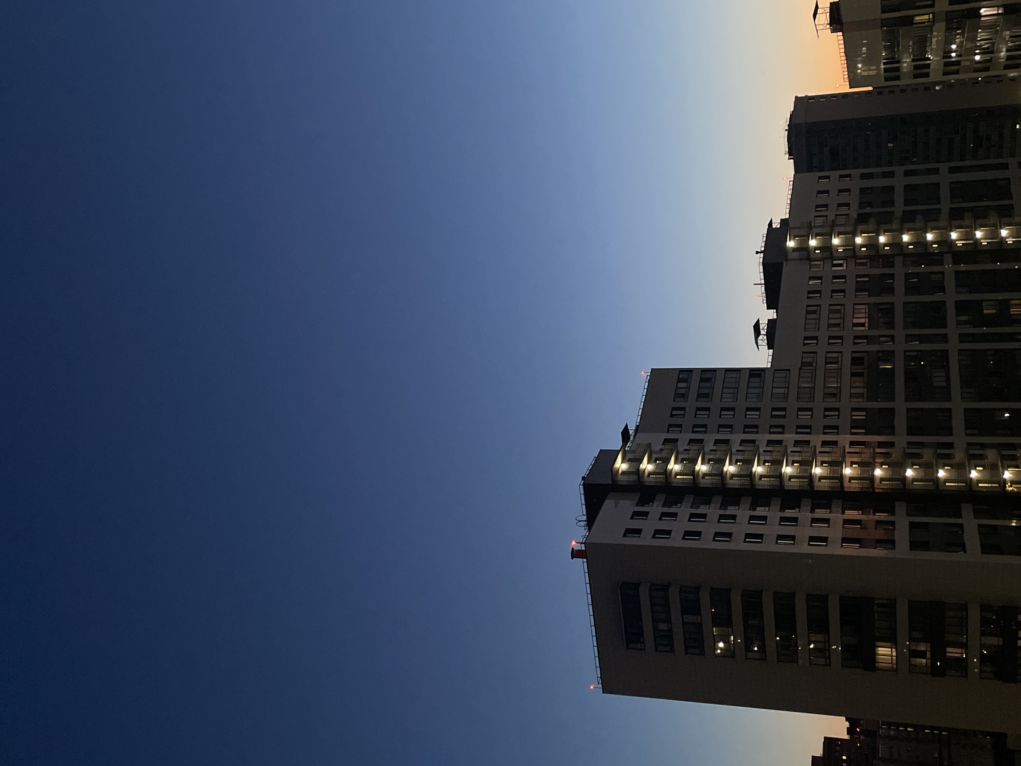 A low-angle, vertical shot captures several residential buildings at dusk in Sinyavino, Russia. The sky transitions from a deep navy blue at the top to a lighter blue and then a faint orange near the horizon on the right. The buildings are modern, multi-story structures with many windows. Some windows are lit, suggesting occupancy. The main building in the foreground is light-colored with dark window frames and balconies. Two smaller red lights are visible on the rooftop of this building, along with what appears to be a red chimney. To the right, other buildings are visible, some with similar lighting and architectural styles. The overall impression is one of an urban landscape during twilight. There are no people visible in the image.