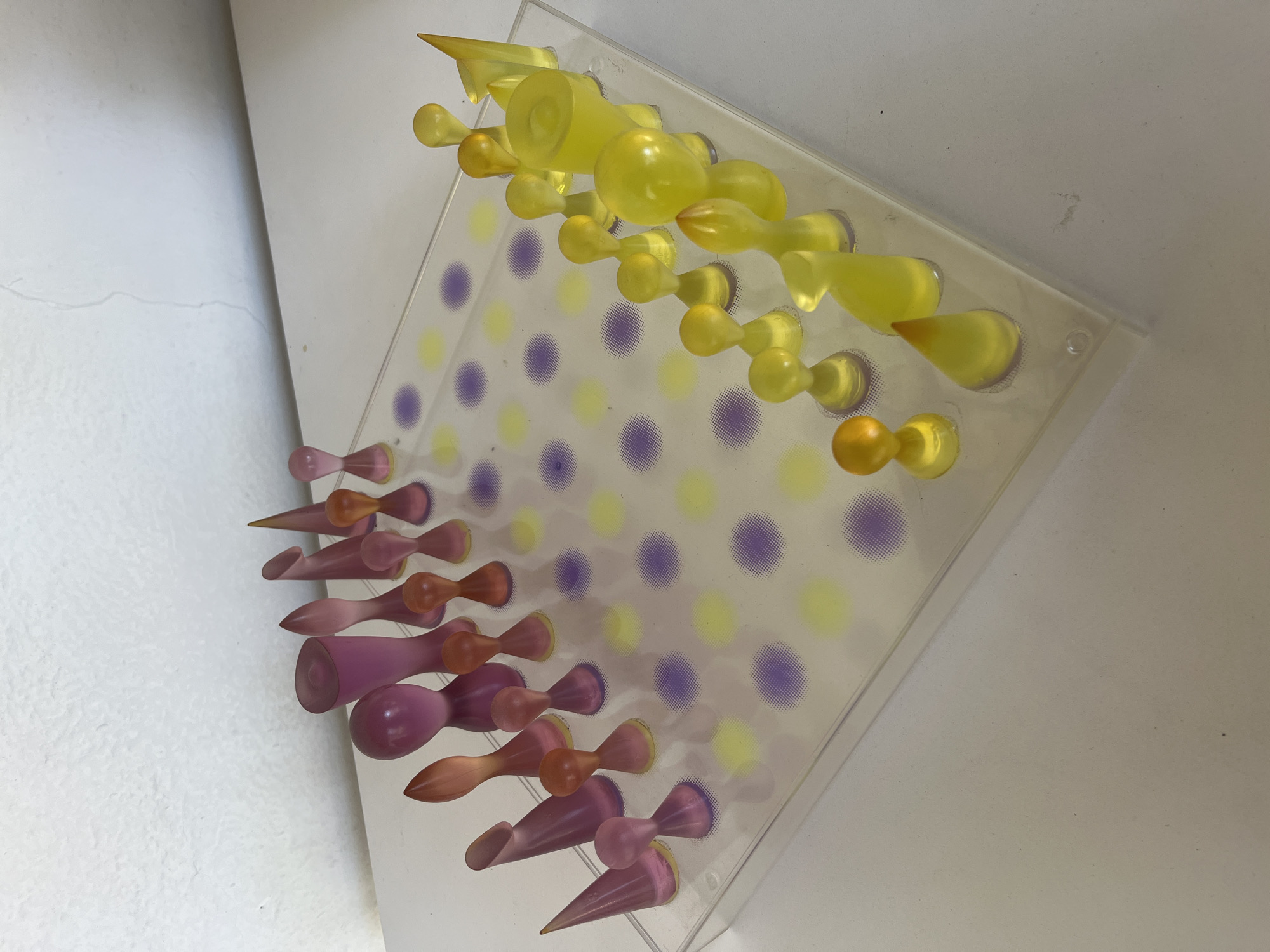 An abstract display piece, likely made of clear acrylic, is shown at an angled perspective. The clear board has a grid pattern of stippled dots in alternating yellow and purple colors. Numerous translucent, sculptural pieces are inserted into the board, protruding outwards.
On the left side of the board, a cluster of pieces in shades of pink and purple extends horizontally. These pieces feature diverse forms, including pointed conical shapes, rounded bulbs, and elongated hourglass-like figures. Many exhibit a gradient of color, appearing darker at their base where they meet the board.
Along the top edge of the board, a similar arrangement of pieces is visible, but these are primarily in shades of yellow and orange. Their shapes also vary, including teardrops, more pronounced conical forms, and rounded, bulbous elements.
The background to the left of the board is a plain white wall with subtle texture and a faint horizontal line. The background to the right and below the board is also a light, plain surface, suggesting it's displayed in an indoor setting against walls or a similar structure. The lighting is bright, enhancing the translucent quality of the colorful pieces. No text is visible in the image.