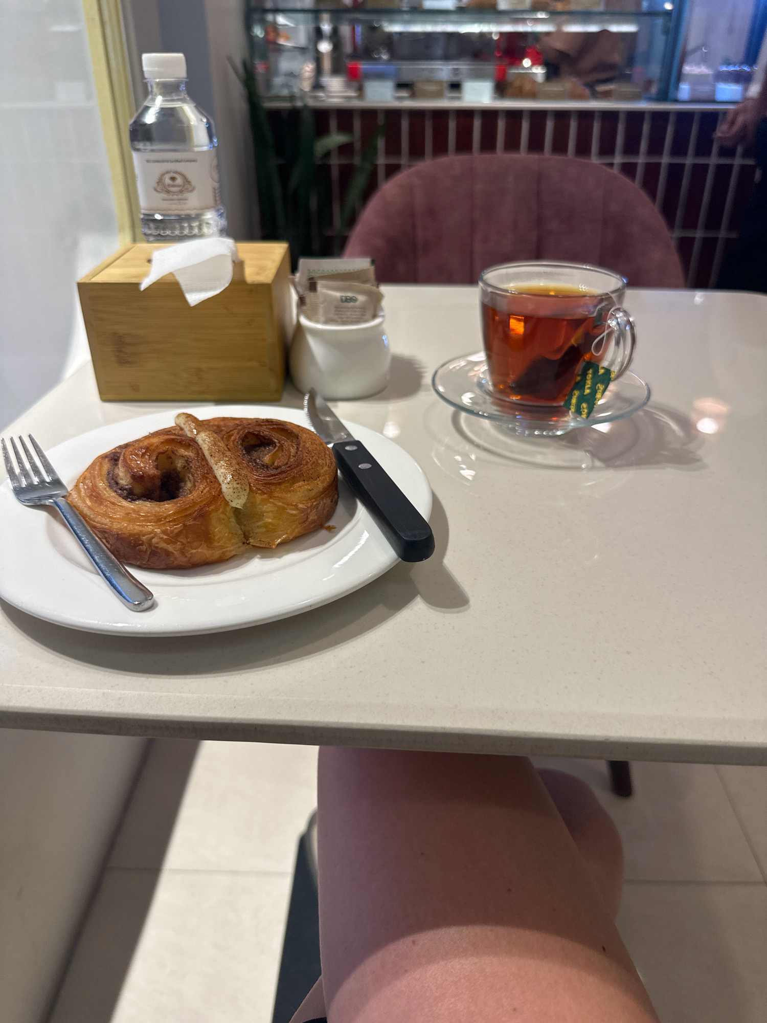 A close-up, eye-level shot captures a table setting in what appears to be a cafe or restaurant. The foreground is dominated by a white plate holding a swirl-shaped pastry, likely a cinnamon roll or similar baked good. A fork rests to the left of the plate, and a knife with a black handle is positioned to the right. Behind the plate, a clear glass mug filled with amber-colored tea sits on a glass saucer. A tea bag tag, green with yellow text, dangles from the mug. To the left of the pastry, a small wooden box dispenses a white tissue. Next to the tissue box, a small white container holds several packets, possibly sugar or creamer. A partially visible water bottle with a white cap stands behind the tissue box. The table surface is smooth and light-colored. In the background, out of focus, is a pink upholstered chair and shelves stocked with various items, suggesting a commercial establishment. The scene is lit by ambient light, creating soft shadows. The setting is a cafe in Kathmandu Metropolitan City, Nepal, where someone is about to enjoy a pastry and tea.